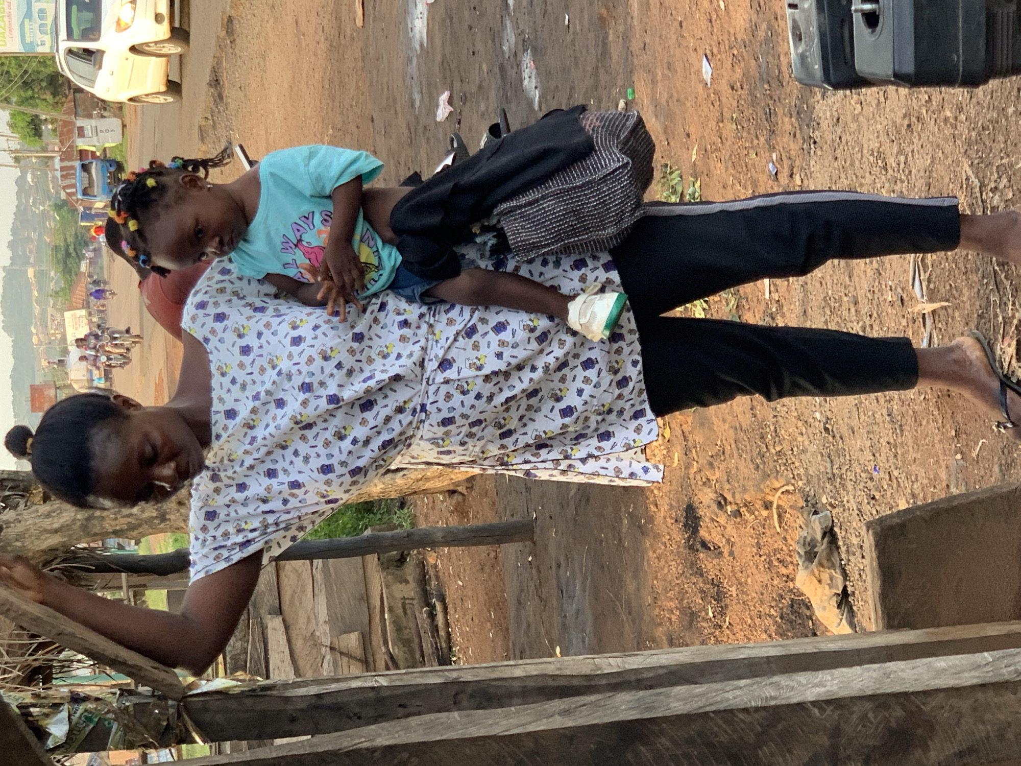 A woman is standing near a wooden structure with a child on her back in Gbeleko, Nigeria. The woman has dark hair pulled back in a bun, wearing black pants with a white stripe, a patterned white top, and flip-flops. The child has dark hair in braids with colorful beads, wearing a blue shirt and jean shorts. A patterned bag is on the woman's back next to the child. The background includes a dirt ground, parts of a building, and a distant view of the city.