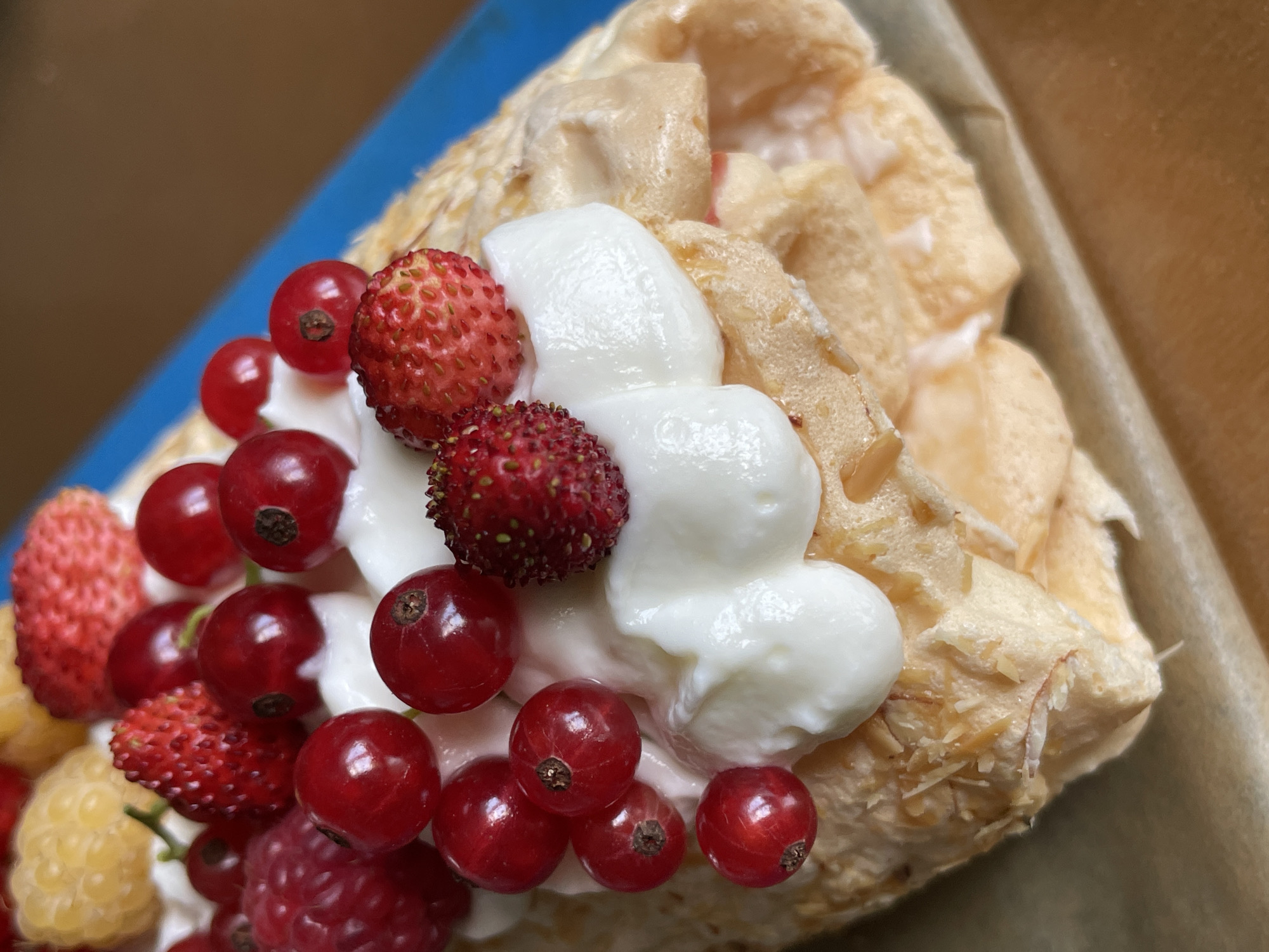 A close-up, top-down shot of a dessert is featured, likely a roulade cake or meringue roll. The dessert is generously topped with dollops of white cream and a colorful assortment of berries including raspberries, red currants, wild strawberries, and yellow raspberries. The cake base appears to be a light, airy meringue or sponge cake, possibly with some sliced almonds sprinkled on the outside. The dessert is presented on a blue platter, with a wooden surface visible in the background, suggesting an indoor setting. The image is well-lit, highlighting the textures and vibrant colors of the fruit and cream. There are no people or animals visible in the image. The scene does not provide any information about the city of Дресвяново, Russia, or any specific location within it.