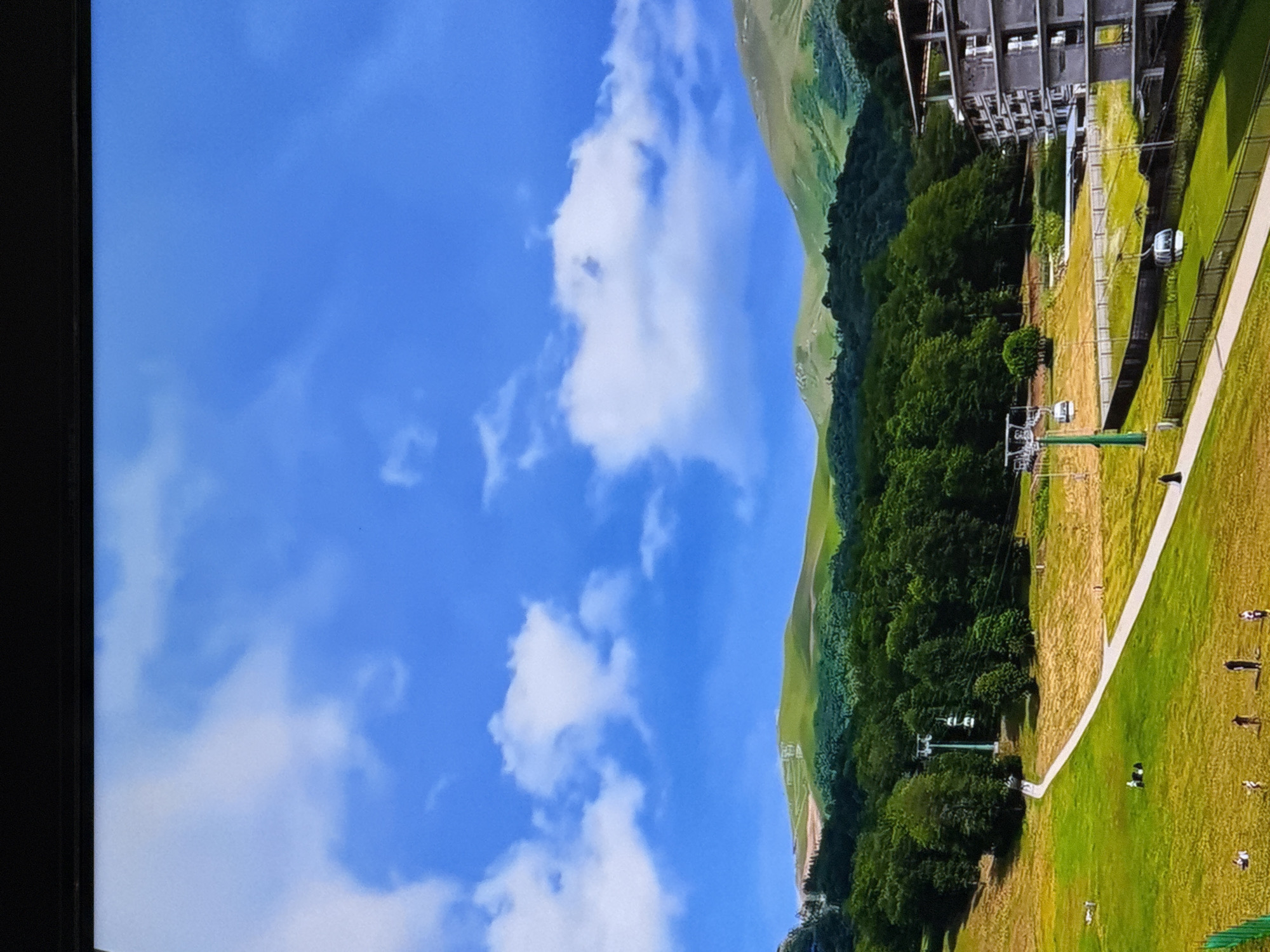 An aerial view captures a lush green mountain landscape with a clear blue sky dotted with white clouds. In the mid-ground, a dense forest of green trees is visible, with a winding path traversing a grassy slope. A ski lift with two cabins is present on the left side of the image, and a modern multi-story building with a gray facade stands on the right. Several small figures are scattered across the grassy area, some appearing to be picnicking or relaxing. A path, possibly leading to the ski lift, is visible in the foreground. The overall scene depicts a scenic mountain resort during a warmer season.