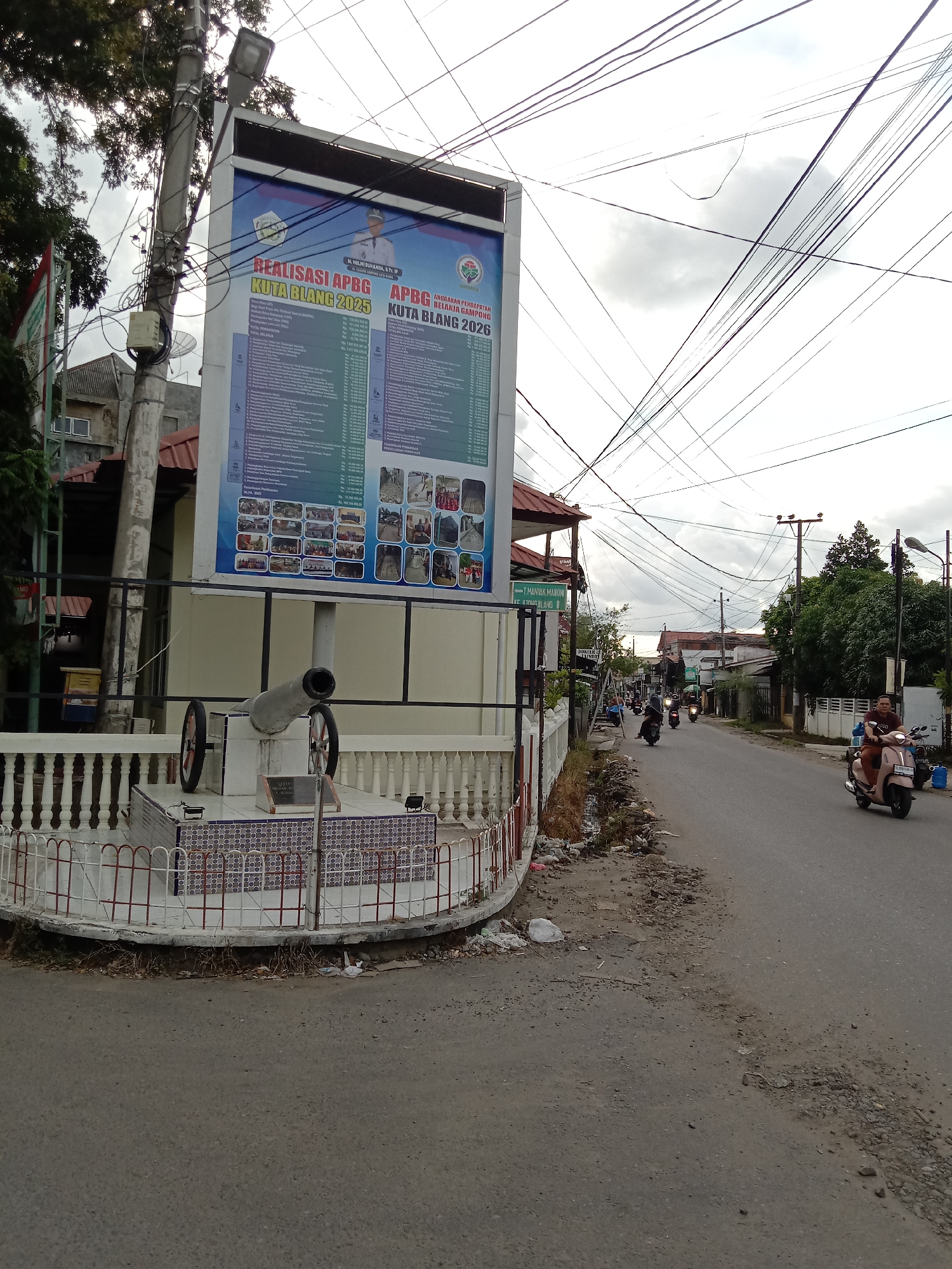 The image depicts a street scene in Kuta Blang, Lhokseumawe, Indonesia. On the left, a large sign displays "Realisasi APBG Kuta Blang 2025" and "APBG Belanja Gampong Kuta Blang 2026," along with images and text related to the village budget. A cannon replica stands in front of the sign, surrounded by a white, decorative fence. The street shows signs of disrepair, with uneven surfaces and debris along the side. Motorcyclists travel along the road, including a person riding a light brown scooter carrying a passenger. Overhead, a complex network of electrical wires crisscrosses the sky. Buildings and trees line the street, contributing to the overall urban environment.