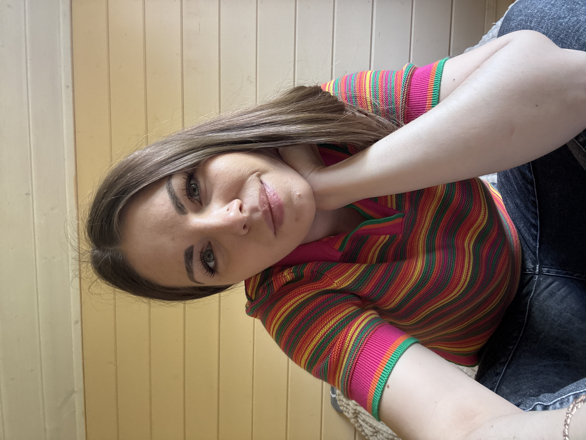 A young woman with shoulder-length brown hair rests her head on her hand. She is wearing a colorful, striped, long-sleeved shirt with pink, green, red, and yellow stripes. She is also wearing dark-colored jeans. The background appears to be a wall with light-colored vertical panels. The location is unknown and cannot be determined from the image.