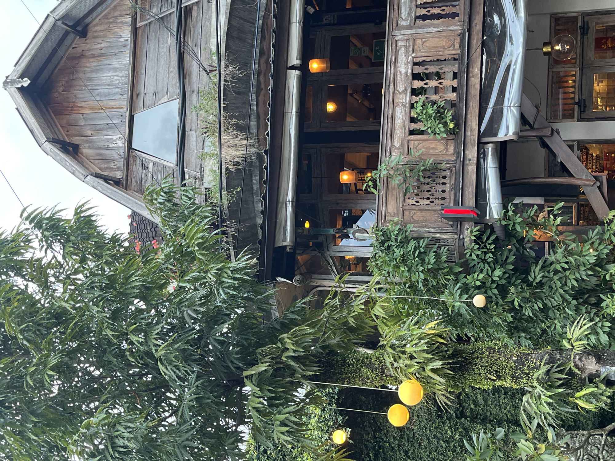 The image depicts a two-story wooden structure in Ubud District, Indonesia, possibly a shop or restaurant. The building has a weathered, rustic aesthetic with visible wood grain and detailed carving. It includes a pitched roof section, a covered awning, and a set of windows illuminated with warm-toned lights. The building's design incorporates various wooden elements with intricate cutouts. A tree and other greenery obscure parts of the structure. Yellow string lights are strung across the branches.