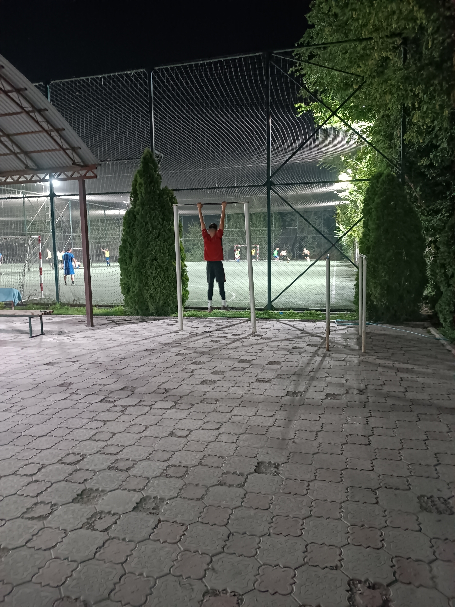 A young male, appearing to be in his late teens or early twenties, is performing a pull-up on a horizontal bar outdoors at night. He is wearing a red t-shirt and dark shorts. The bar is supported by two vertical posts. The ground in the foreground is paved with interlocking stones. In the mid-ground, a soccer field is visible with a net enclosure and lighting. Several other individuals are present on the soccer field, some appearing to be playing soccer. Two tall, green evergreen trees are situated behind the pull-up bar. To the left, a covered seating area with a corrugated metal roof is present. The scene is illuminated by artificial lights.