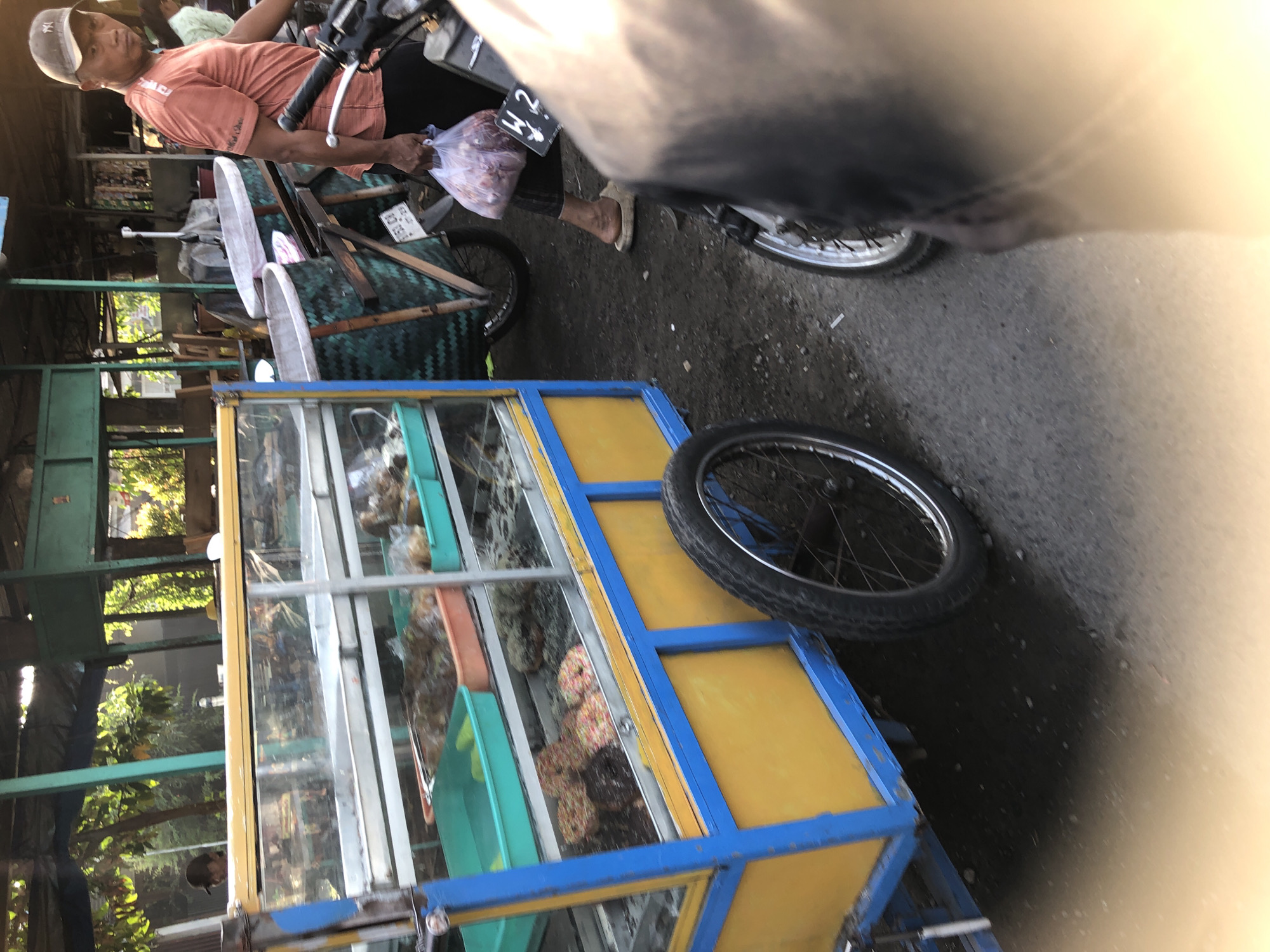 The image depicts a street scene in Balongdowo, Indonesia, with a focus on a food cart and its surroundings. In the foreground, a person wearing an orange t-shirt and a cap is visible from the waist down, holding a plastic bag filled with food. They are standing near a motorcycle. To the left, a food cart with a yellow and blue frame is parked. The cart has glass display shelves filled with various food items, including what appear to be donuts and other snacks. Behind the food cart, two large woven baskets are attached to a bicycle or cart. The background shows a shaded area with trees and some structures that suggest a market or roadside food stalls. The overall scene conveys a typical Indonesian street market environment.