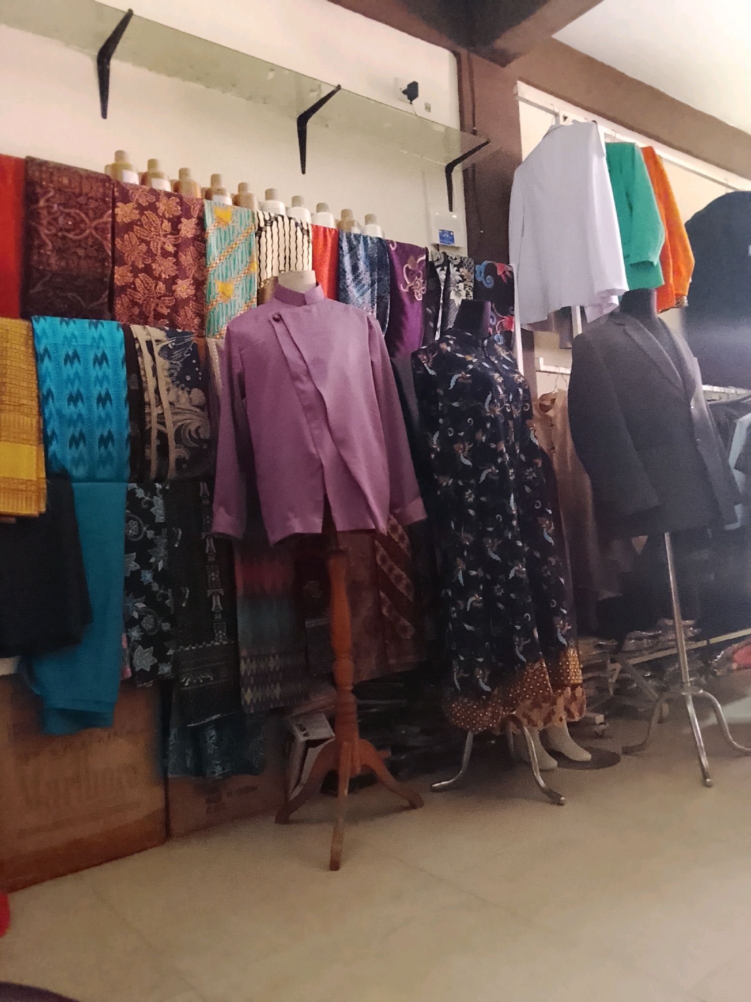 The image depicts a retail store displaying various textiles and clothing, likely a batik shop. The scene is organized with fabrics folded and hung on display racks and mannequins. In the foreground, on the left, are folded fabric swatches in vibrant colors and patterns, including a bright yellow piece and a blue patterned fabric. To the right, a mannequin is dressed in a light purple long-sleeved shirt with a unique asymmetrical closure, displayed on a wooden stand. Next to it, another mannequin wears a long, dark blue dress adorned with a delicate white and orange floral pattern, with the hem featuring a patterned border. Behind these figures, more fabrics are visible, including patterned batiks in shades of brown and red. On the upper right side of the image, several shirts are hung on a rack: a white shirt, a bright green shirt, and an orange shirt. A dark suit jacket is also displayed on a mannequin to the right of the floral dress. The store appears to have a tiled floor and a suspended ceiling with wooden beams. The overall impression is a well-stocked establishment offering traditional Indonesian textiles and garments. This scene is consistent with shops found in the city of Tanjung Balai Karimun, Indonesia, which is known for its textile trade, including batik.