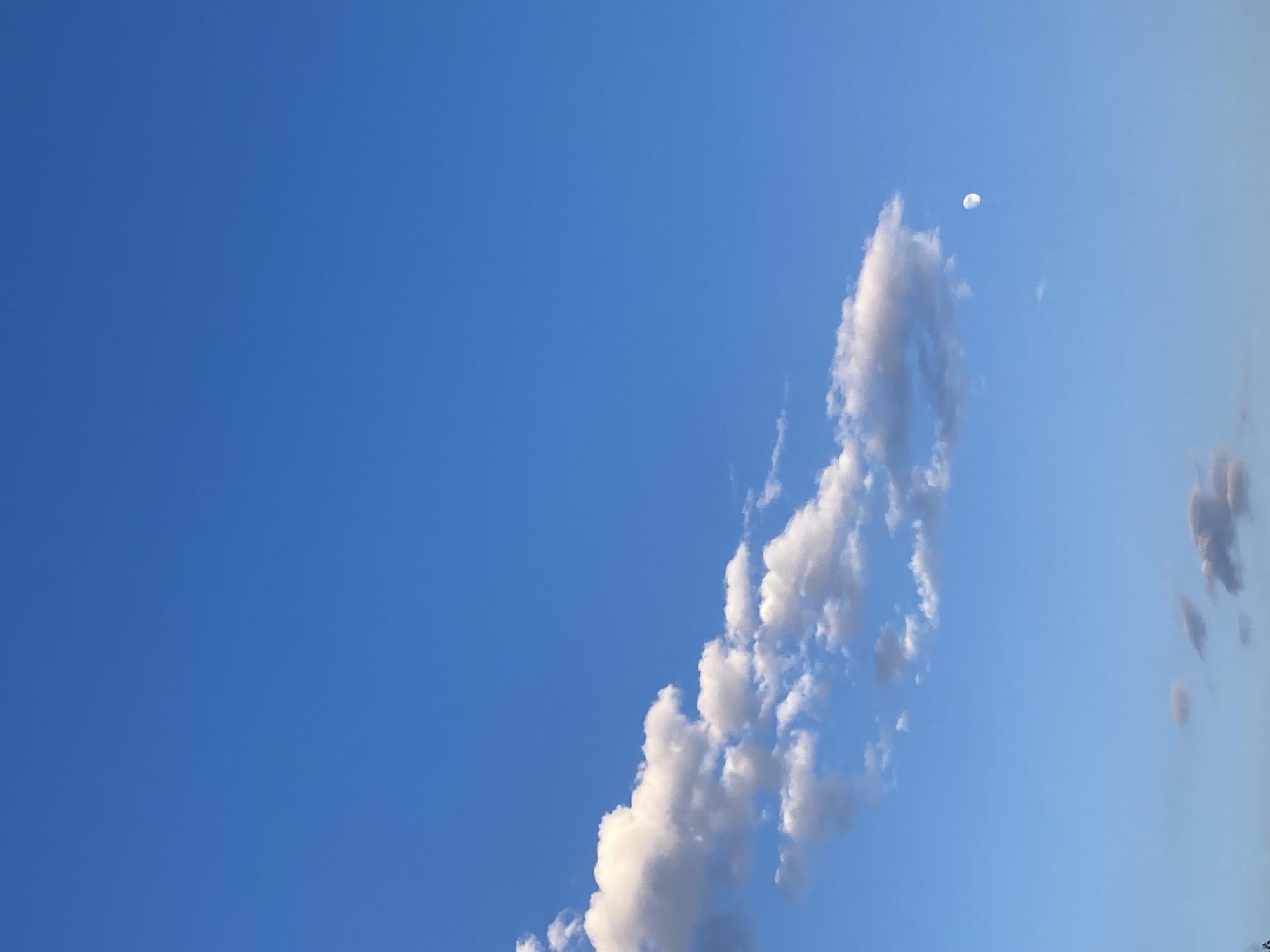 The image shows a clear blue sky with scattered clouds and a visible crescent moon. The clouds are white and fluffy in the upper left portion of the frame, with a few darker, smaller clouds towards the bottom. The moon is a small, luminous crescent on the right side of the image. The scene appears to be outdoors, likely during the daytime given the bright blue sky, though the presence of the moon suggests it could be late afternoon or early evening. There are no people or structures visible in the frame, creating a minimalist and atmospheric depiction of the sky. The specific location of Avanos İlçe Merkezi, Turkey, is not visually evident from the provided image.