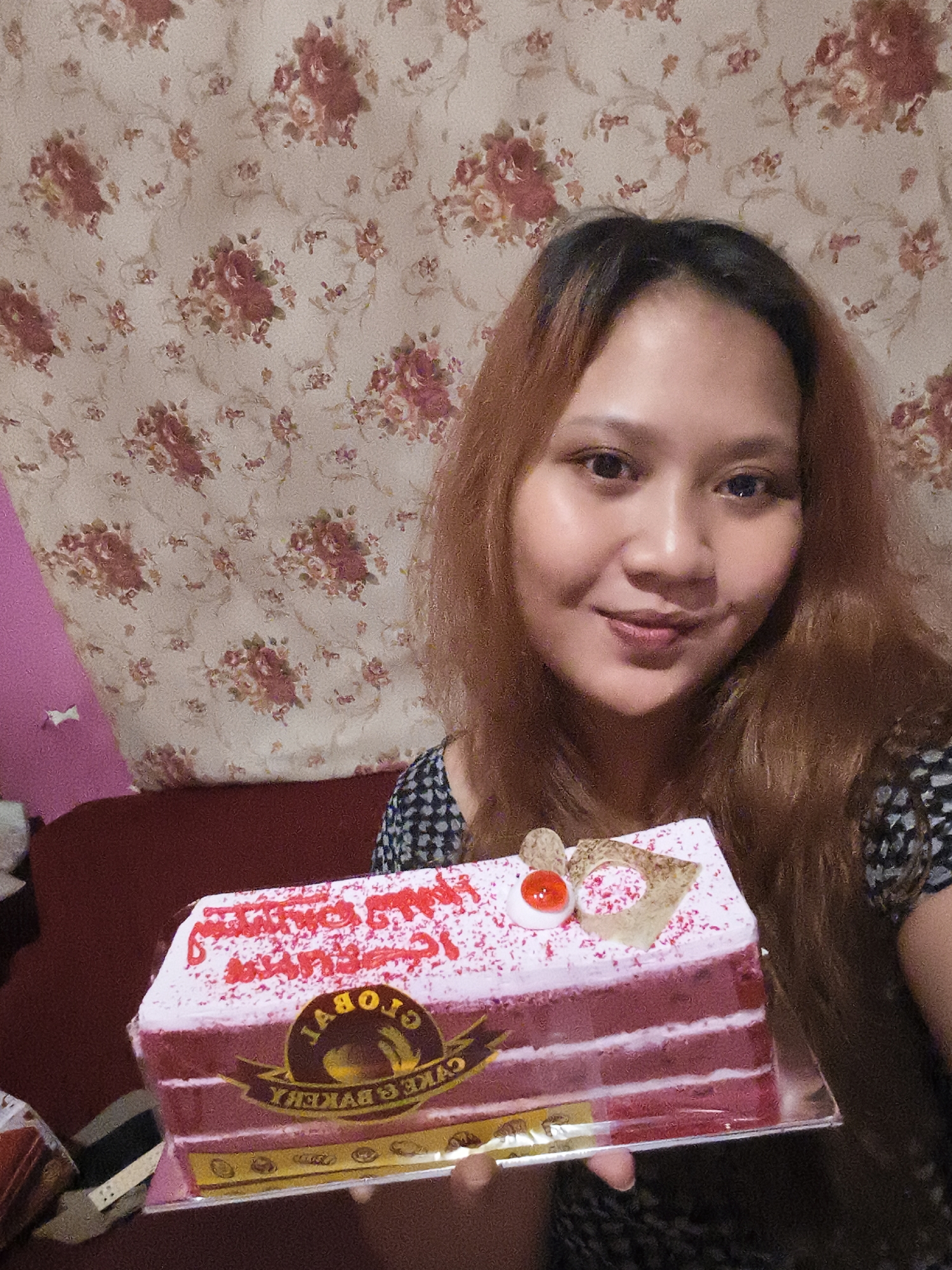 A woman with long, auburn hair and fair skin is holding a rectangular, red velvet cake with white frosting. The cake is decorated with red sprinkles and red cursive writing, possibly a birthday message. A circular logo with the word "GLOBAL" is visible on the side of the cake. The woman is smiling faintly at the camera. She is wearing a dark, patterned top. The background features a floral patterned curtain and a hint of a maroon sofa. The scene appears to be indoors, likely in a home in Depok, Indonesia.