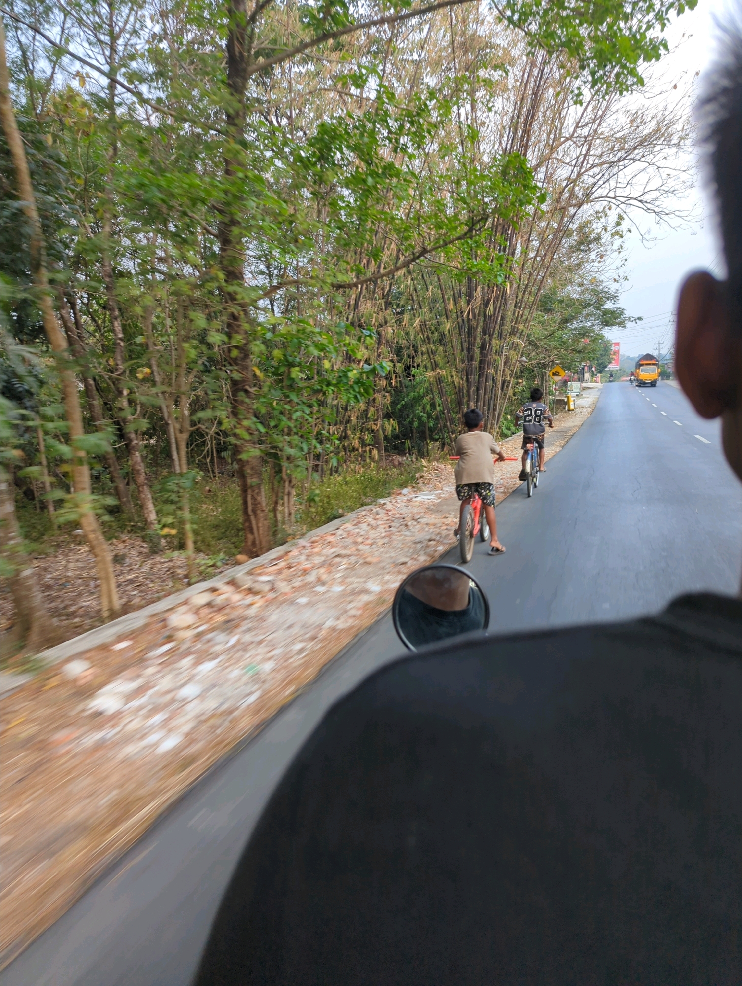 Two young boys are riding bicycles on a paved road lined with trees and bamboo. The boy in the foreground is wearing a light brown shirt and patterned shorts, riding a red bicycle. The boy behind him is wearing a black and white shirt with the number 28 on it, riding a blue bicycle. A yellow truck is visible in the distance on the road. The scene appears to be a rural area in Senggrong, Indonesia, with lush greenery and a clear sky. The image is taken from behind the viewer, looking forward over the shoulder of someone on a vehicle, with a rearview mirror visible in the foreground.