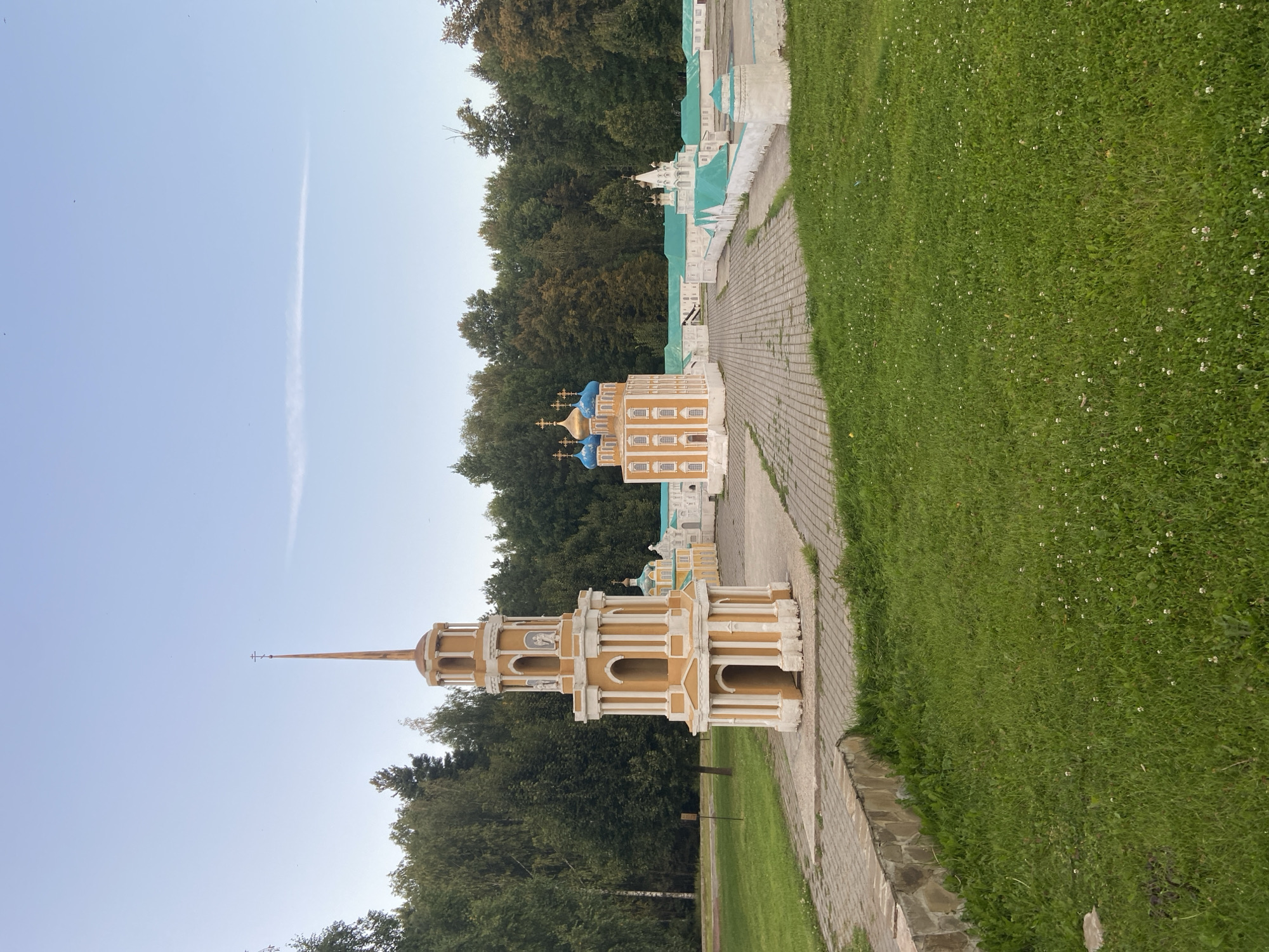 This is a horizontal, eye-level, medium shot of a miniature park with various church models. In the foreground on the left, a tall, yellow and white bell tower stands prominently. Its tiered structure features arched openings and decorative columns. To the right of the bell tower, a larger church model with blue domes and yellow walls is visible, followed by a series of white buildings with teal roofs, forming a courtyard. The models are situated on a paved area surrounded by lush green grass and a dense line of trees in the background. The sky is a clear, pale blue with a few wispy clouds and several small, dark specks that might be birds. The overall scene depicts a peaceful, sunny day in an outdoor recreational area. The location is identified as Chulis, Russia. There are no people or animals present in the image.