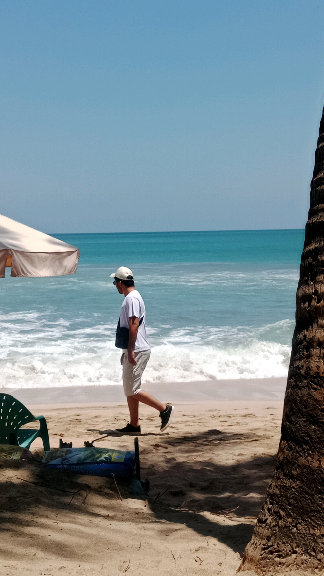 A man wearing a white t-shirt, beige shorts, a white cap, sunglasses, and black shoes walks along the beach in Kuta, Indonesia. He is carrying a dark messenger bag over his left shoulder. In the background, the ocean has light blue water with white waves breaking on the shore. The sky is a clear, light blue. In the foreground, a green plastic beach chair sits on the sand to the left, and a blue object, possibly a bag or a cooler, is partially covered by sand and sticks to the right of the chair. A portion of a palm tree trunk is visible on the far right edge of the image. A beige beach umbrella is partially visible in the upper left corner.
