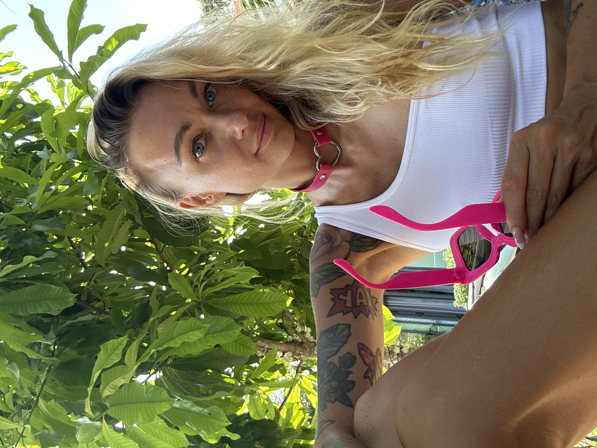In Mui Ne, Vietnam, a blonde woman with blue eyes and tattoos on her arm poses for a photo in front of lush green foliage. She is wearing a white ribbed tank top and a pink choker with a heart-shaped pendant. In her right hand, she holds a pair of pink cat-eye sunglasses. The sunlight filters through the leaves, creating a bright and airy atmosphere. The woman is looking directly at the camera with a slight smile.