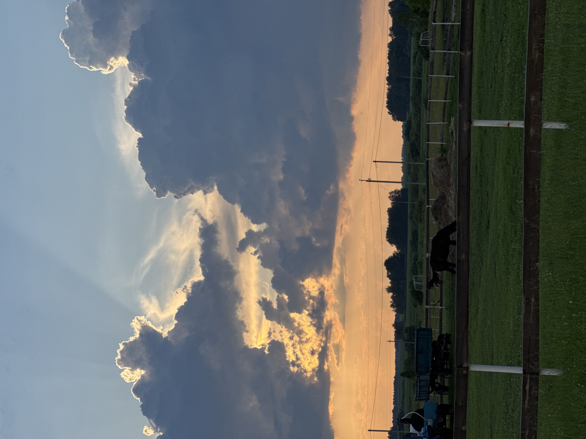 This media file captures a rural scene in the village of Bigila, Russia, during what appears to be late afternoon or early evening. The sky is dramatic, dominated by large, dark clouds illuminated by the setting sun, which casts a warm, golden glow through breaks in the clouds and along the horizon. Rays of sunlight fan out from behind the clouds, creating a dramatic effect.
The foreground features a grassy field enclosed by a wooden fence, typical of a farm or pasture. A black horse is visible in the field, standing near the fence. Further back, several agricultural vehicles, including a trailer and a tractor, are parked on the grass. A bale of hay is also present. Beyond the field, a line of trees marks the edge of the landscape. Telephone poles and power lines are visible against the colorful sky, indicating the presence of civilization.
The overall mood is one of natural beauty and tranquility, with the dramatic sky adding a sense of awe. The time of day is suggested by the quality of light and the colors in the sky, and the location cues point to a rural setting in Russia. No text is visible in the image.