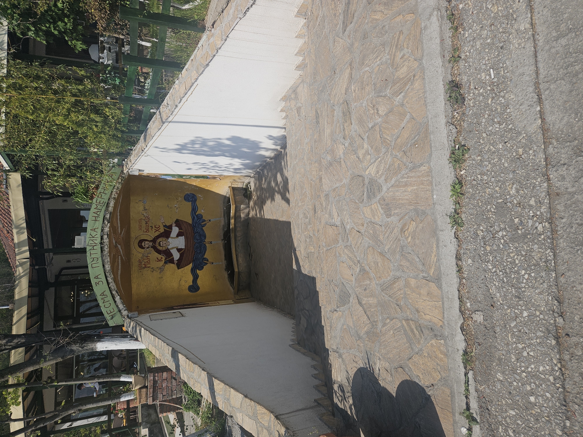 This image captures an outdoor scene in Velika Plana Municipality, Serbia, showing a stone-paved pathway leading down towards an arched entrance. The scene is brightly lit by the sun, casting distinct shadows on the ground and walls, suggesting a clear day in the morning or afternoon.
The foreground features a textured stone pathway on the right, made of irregularly shaped light brown stones. Adjacent to this, on the left, is a concrete border separating it from a darker, asphalt-like surface where small weeds grow in the cracks. A discarded cigarette butt is visible on the darker surface.
The focal point is a white-walled structure with a curved archway, adorned with a vibrant yellow background and religious iconography. This artwork depicts two haloed figures, presumably saints, in a boat sailing on stylized blue waves. Cyrillic text identifies the left figure as "СВЕТИ ПЕТАР" (Saint Peter) and the right figure, holding a book, as "СВЕТИ ЈОВАН БОГОСЛОВ ХС" (Saint John the Theologian HS/Christ). Above the arch, a green band carries the Cyrillic inscription "ЧЕСМА ЗА ПУТНИКА НАМЕРНИКА," which translates to "FOUNTAIN FOR TRAVELERS / WAYFARERS."
Beyond the archway, obscured by dense green foliage and trees, parts of a building with large windows and dark framing are visible, suggesting a restaurant, cafe, or guesthouse. A green wooden trellis or fence can be seen on the far left, adding to the garden-like atmosphere. No people are visible, and the scene is static.