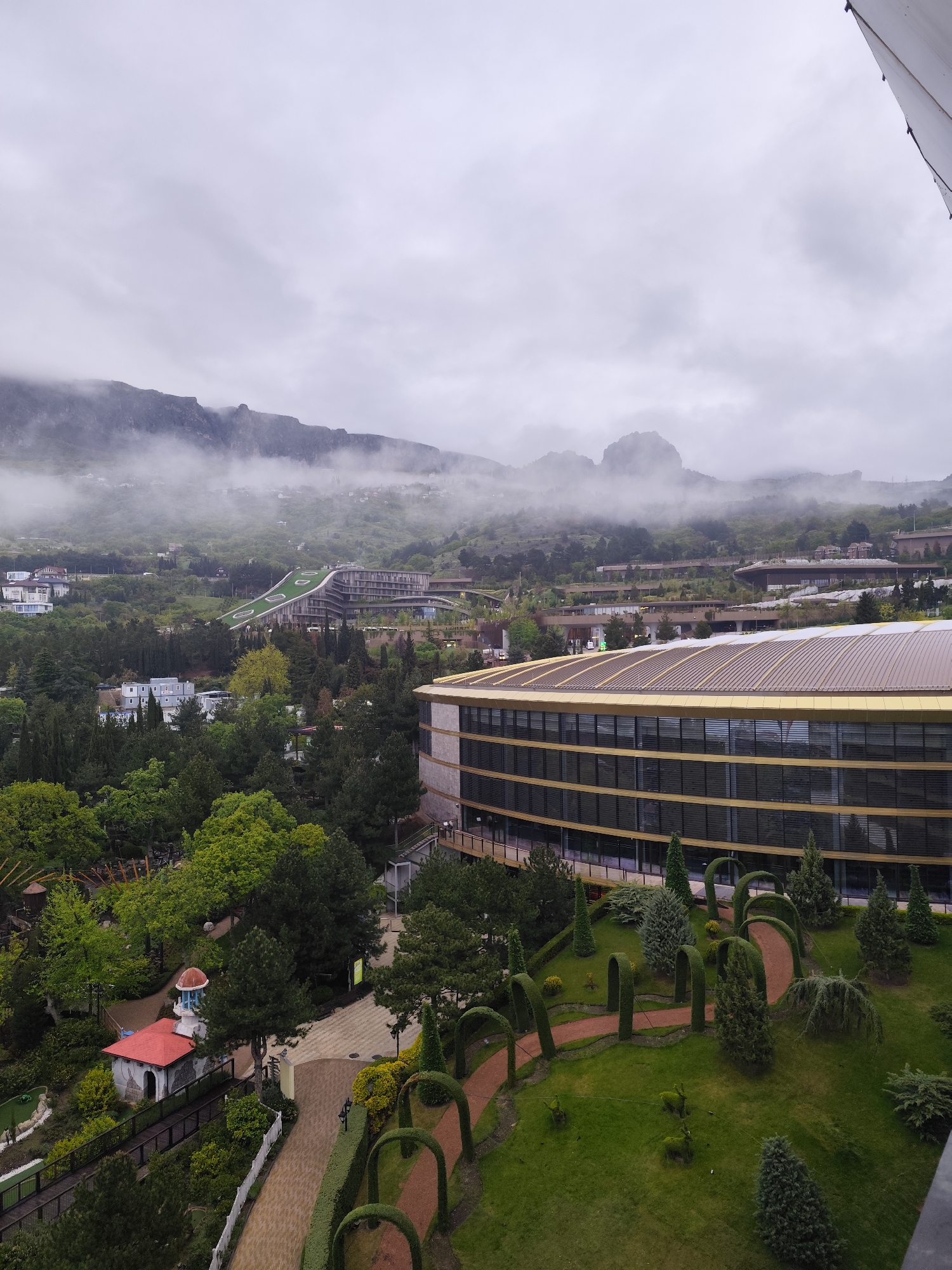 The image depicts a cityscape in Симеизский поселковый совет, Ukraine, under an overcast sky. Mountains shrouded in mist dominate the background. In the foreground, a large modern building with a gold-colored roof is visible, and its multi-story facade is predominantly glass. A well-manicured garden with hedges shaped into arches and walking paths extends from the building. A small building with a red roof and a white tower sits among trees in the middle ground. The general atmosphere is subdued due to the weather, with the greenery providing a contrast to the grayness of the sky and building.