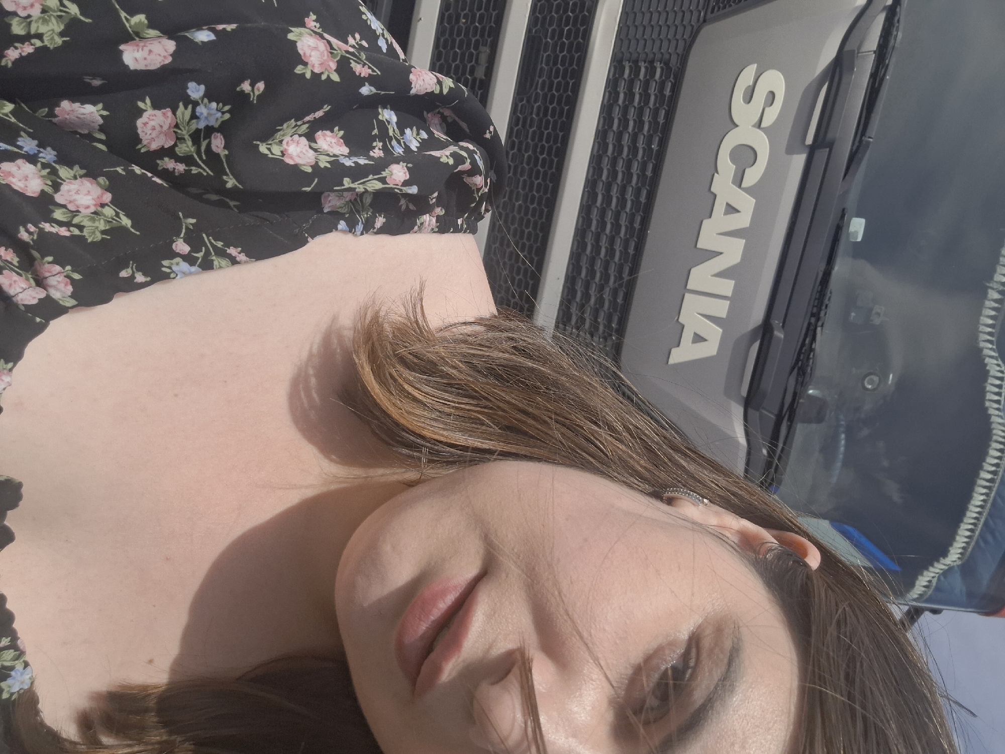 A woman is angled in the foreground, and a Scania truck is in the background. The woman has light skin, brown hair, and is wearing a black floral top. The Scania truck is gray, with the company name "SCANIA" prominently displayed in silver lettering. The setting appears to be outdoors with bright lighting, but it's unknown if the location is Izhevsk, Russia.