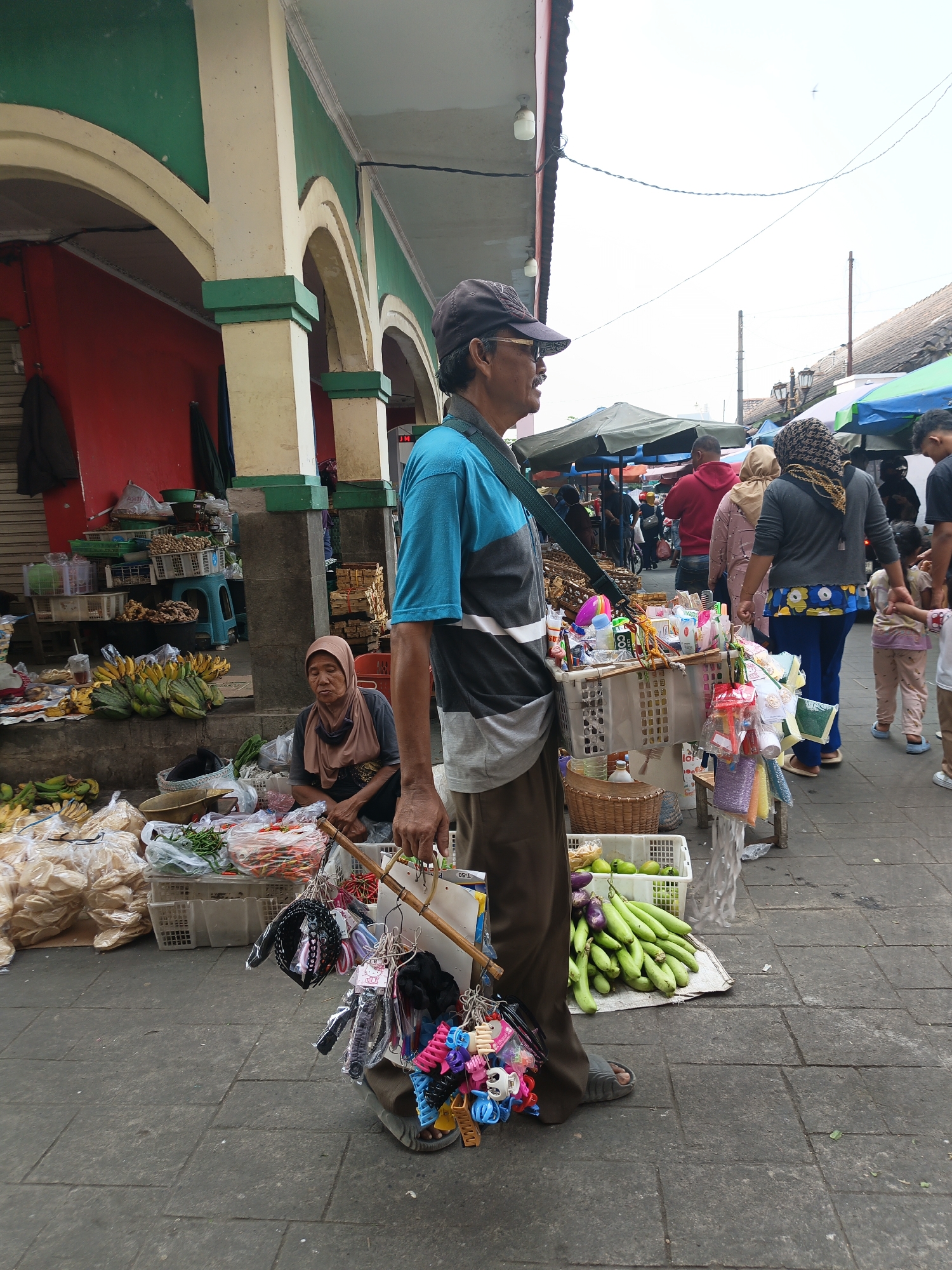 The image captures a street scene in Klaten, Indonesia, likely a market or roadside stall area. In the foreground, a man wearing a blue and grey t-shirt, brown pants, a cap, and glasses stands on a tiled sidewalk, holding a wooden pole from which various colorful hair accessories and small toys are suspended. To his left, a woman wearing a headscarf sits on the ground beside a display of green vegetables and other goods. Behind them, a stall with arches and a red wall is visible, suggesting a more permanent structure. Further in the background, people are browsing other stalls, some covered by awnings, and the sky is overcast. The ground is paved with rectangular tiles.