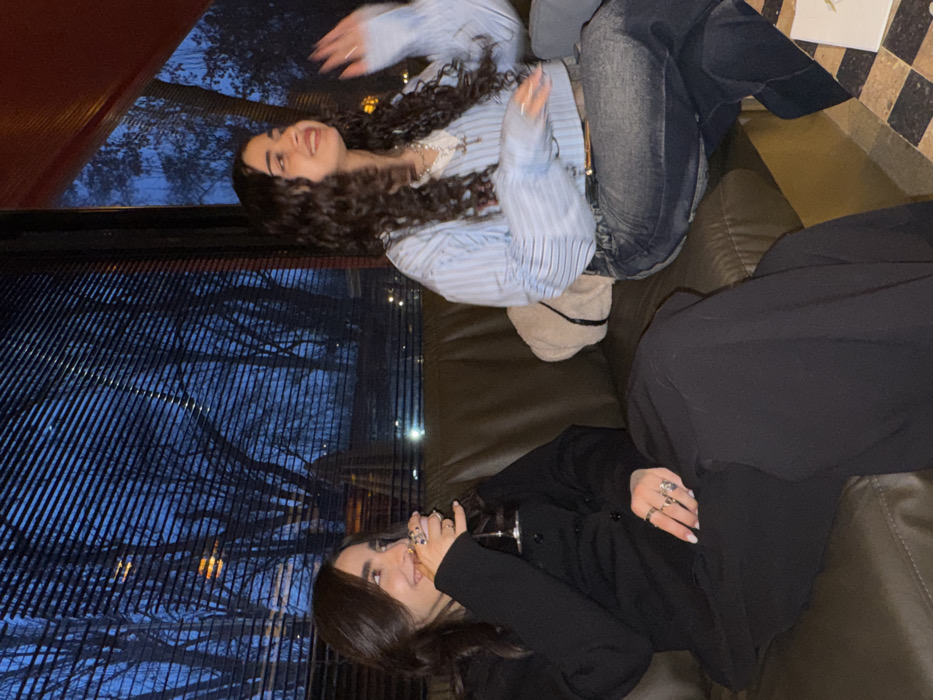 Two young adult women are seated on a dark green upholstered bench inside an establishment in Yerevan, Armenia, during the nighttime hours. The woman on the left has dark brown, shoulder-length hair, wears a black collared top and black trousers, and multiple rings on her left hand. She holds a stemmed glass with a clear liquid in her right hand and is smiling while looking towards her right. The woman on the right has long, dark, curly hair, wears a light blue and white striped cropped shirt over a white underlayer, blue denim jeans, a necklace with a cross pendant, and multiple rings on her right hand. Her right hand is raised and blurred, indicative of motion, and she is also smiling and looking towards her right. A light-colored shoulder bag rests on the bench next to her. Behind the women is a large window with horizontal blinds, revealing a dark outdoor scene with silhouetted bare tree branches against a dark blue sky and distant artificial lights. A corner of a table with a black and white checkered surface is visible in the lower right foreground.