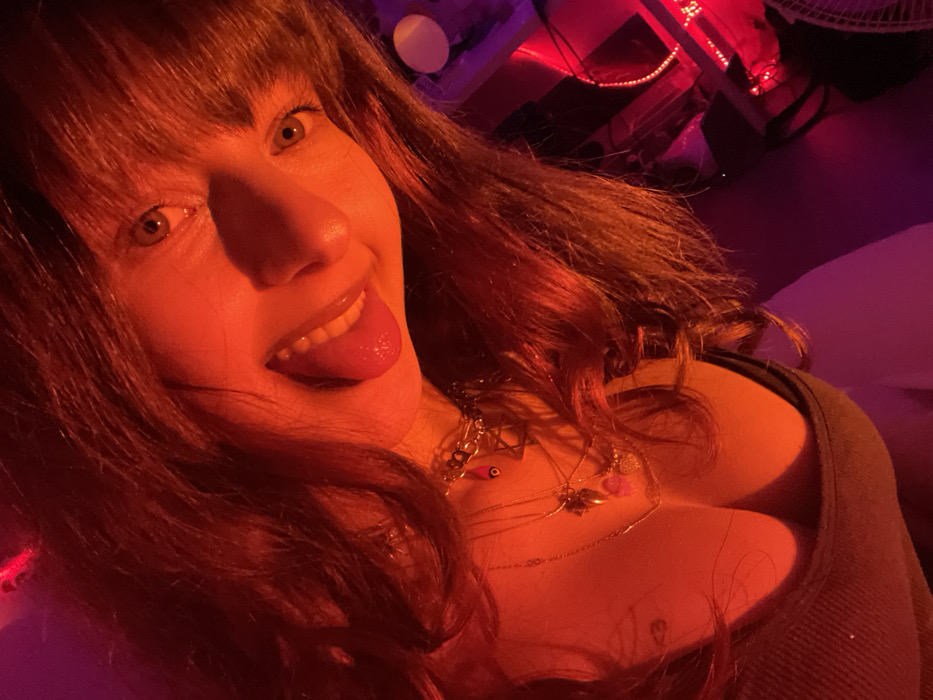 A woman with reddish-brown hair and bangs poses close to the camera. She sticks out her tongue and smiles, displaying her teeth. She wears several necklaces. Red lighting casts a warm glow over the scene. Background elements include a white round object, what appears to be a table with red lights, and part of a fan. The woman's clothing is visible.