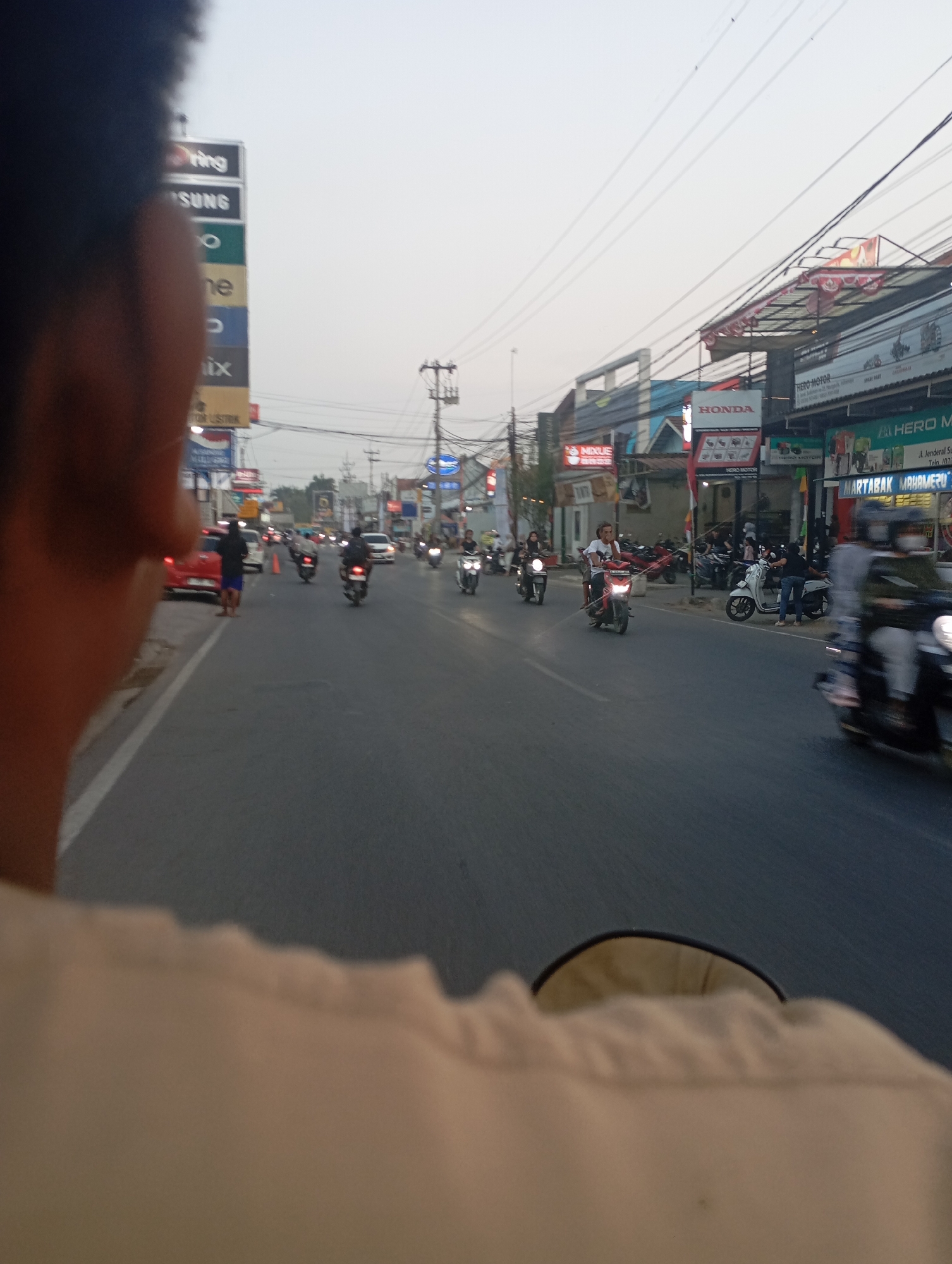 The image depicts a street scene in Kecamatan Haurgeulis, Indonesia. The perspective is from a person riding on the back of a motorcycle, with their shoulder and the back of their head blurred in the foreground. The street is paved and appears to be a main road with multiple lanes. Several motorcycles and a few cars are visible on the road, with most of the motorcycles being ridden by individuals. On the right side of the road, there are buildings that appear to be shops or businesses, some with awnings and signs, including "HONDA" and "HERO MOTOR". A tall billboard structure with multiple brand logos, including "SAMSUNG" and "OPPO", is present on the left side of the road. The sky is overcast or hazy, and the lighting suggests it is late afternoon or early evening. The environment is urban, characterized by traffic, commercial establishments, and street infrastructure.