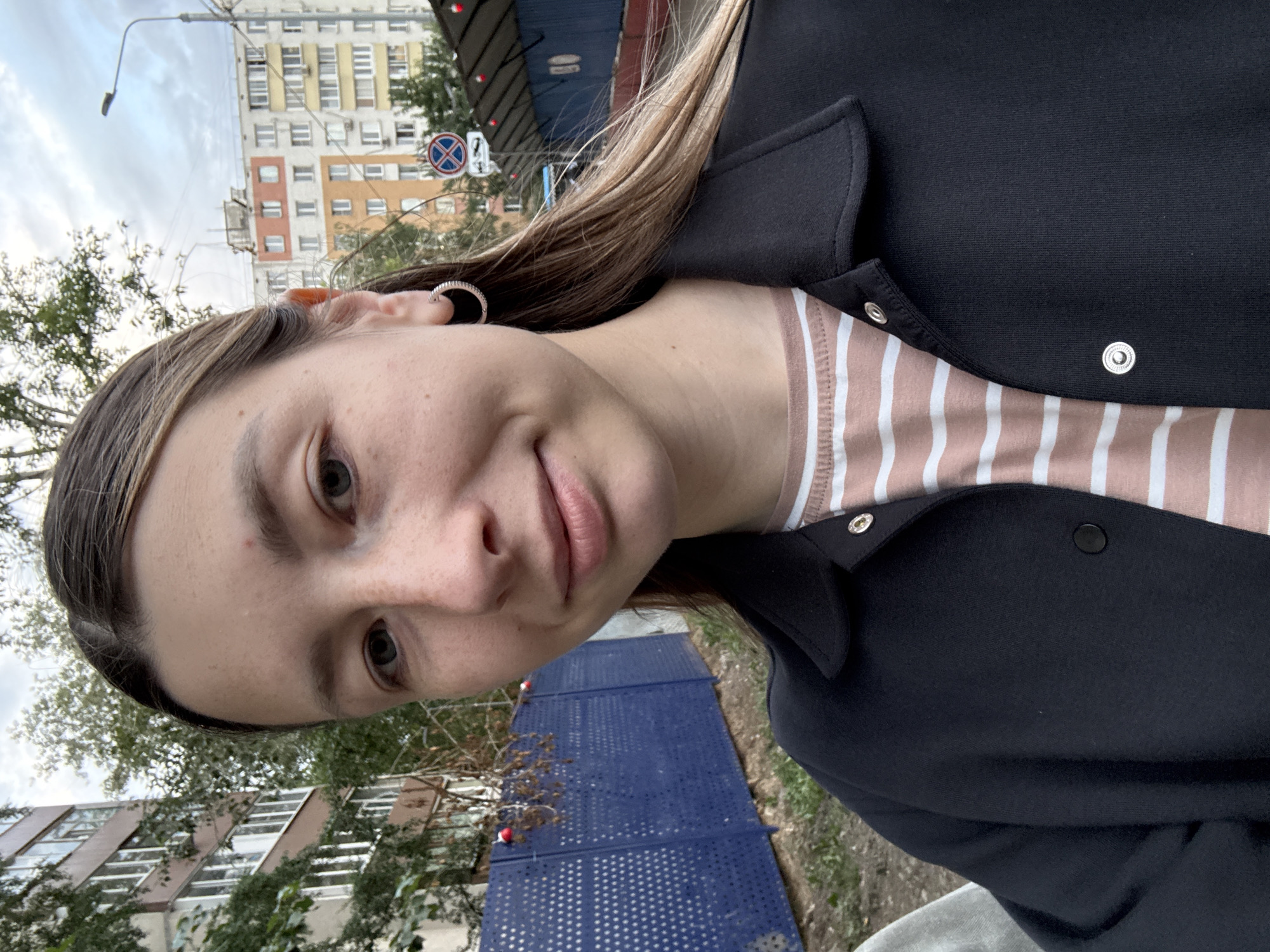 The image shows a young woman, likely in her late teens or early twenties, taking a selfie outdoors. She has light brown hair with blonde highlights, fair skin with freckles, and is wearing a black jacket over a pink and white striped t-shirt. She is also wearing a silver hoop earring in her left ear.
In the background, there are residential buildings typical of Moscow, Russia, with multiple stories and balconies. One building is a light beige color with dark brown window frames and balconies, while another is a multi-colored structure with sections of orange, yellow, and white. There is a blue fence with a perforated pattern behind the woman, and trees with sparse foliage are visible. Further in the background, street signs, including a "no stopping" sign, and a lamppost are present, suggesting an urban setting. The overall environment appears to be overcast, with a cloudy sky. The image is taken from chest up, focusing on the woman's face and upper body.