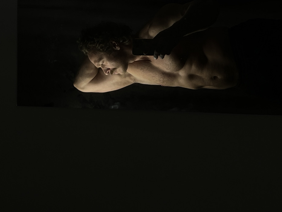 The image depicts an adult male, with a muscular physique, curly dark hair, and a mustache, captured in what appears to be a mirror selfie. He is shirtless, exposing his torso with visible abdominal muscles, and wears dark bottoms, partially obscured from view. The man is positioned in profile, facing to the viewer's right and looking upwards, with his right arm bent and hand placed behind his head. His left hand holds a dark-colored smartphone, partially obscuring the lower portion of his face. Strong, directional lighting from the left illuminates his features and body, creating stark contrast against a predominantly dark background. The scene is indoors. The specific location within Saint Petersburg, Russia, cannot be determined from the visual evidence provided.