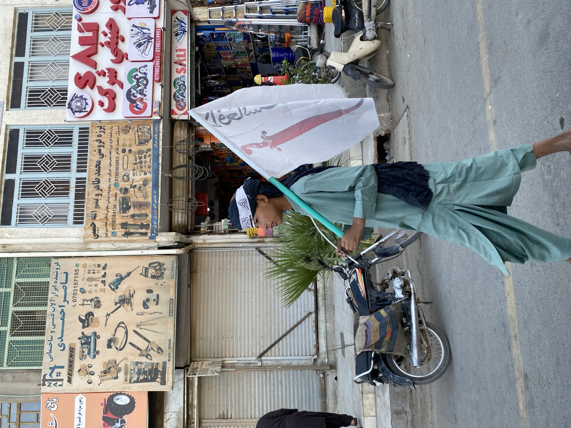 A young male wearing a light green shalwar kameez and a black head covering walks on a paved street in Kandahar, Afghanistan. He carries a white flag with a red graphic and Arabic text. A black motorcycle with a colorful woven seat cover is parked on the side of the street. In the background, storefronts display advertisements for tools and machinery. The buildings have metal roll-down security shutters.