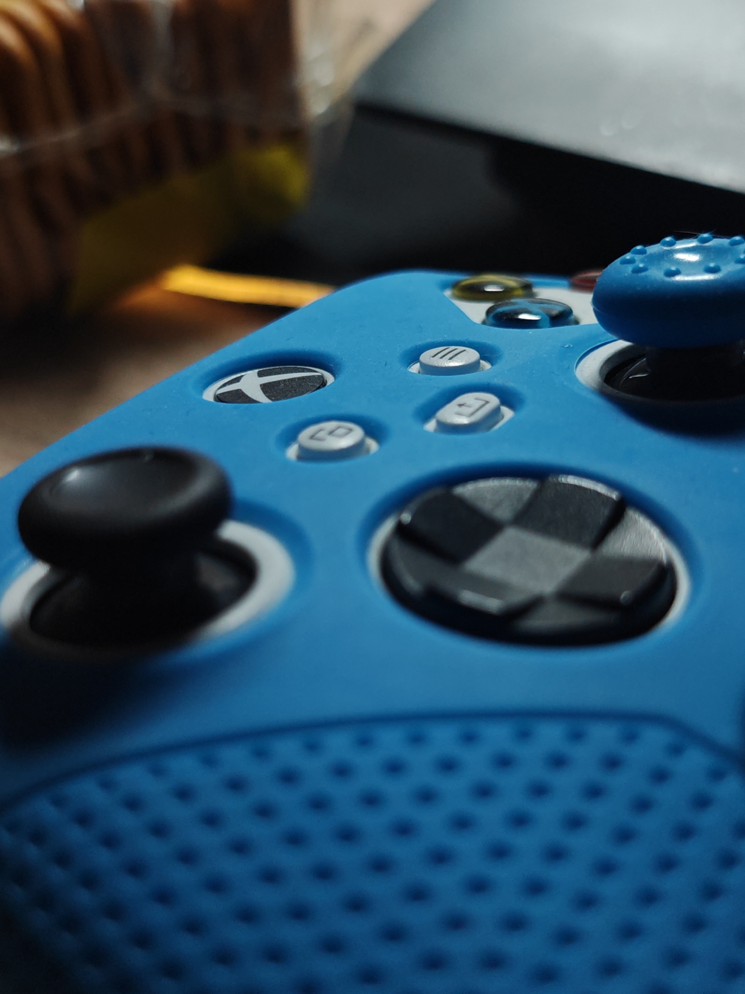 The image shows a close-up of a blue Xbox controller. The focus is on the left side of the controller, highlighting the analog stick, D-pad, and some of the face buttons. A clear protective film seems to be placed over the joystick. The foreground shows the texture of the controller's grip. The background is blurry, but the top left corner reveals a stack of what appears to be crackers or cookies in a package. A dark, indistinct object is in the top right.
The image does not provide information to identify the location in the city of Hrodna, Belarus. It is a generic photograph of a gaming controller that could have been taken anywhere.
