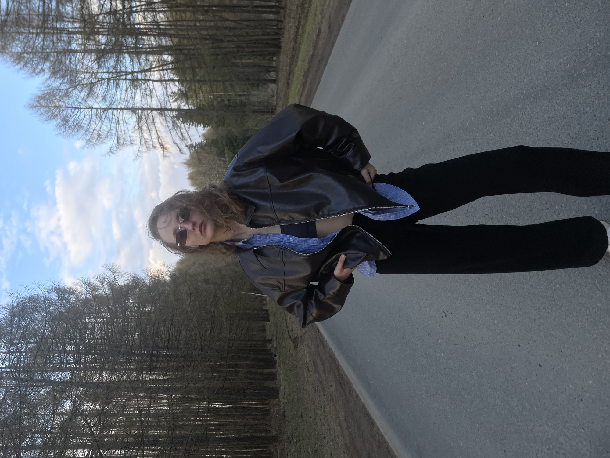 A full-body shot depicts a person standing on an asphalt road in Podshivalovo, Russia. The individual, who appears to be female, has long, wavy brown hair and is wearing dark sunglasses. She is dressed in a black leather jacket, unzipped to reveal a blue and white striped button-down shirt, which is open over a black top or bra. Black pants and light-colored sneakers are also visible. The person stands casually, looking towards the camera, with a confident or relaxed pose.
The road is bordered on both sides by a dense forest of tall, deciduous trees, mostly bare of leaves, suggesting either late autumn, winter, or early spring. The sky above is a light blue with scattered white clouds, indicating clear daytime conditions. The scene captures an outdoor portrait on a rural or semi-rural road amidst nature. There is no visible text in the image.