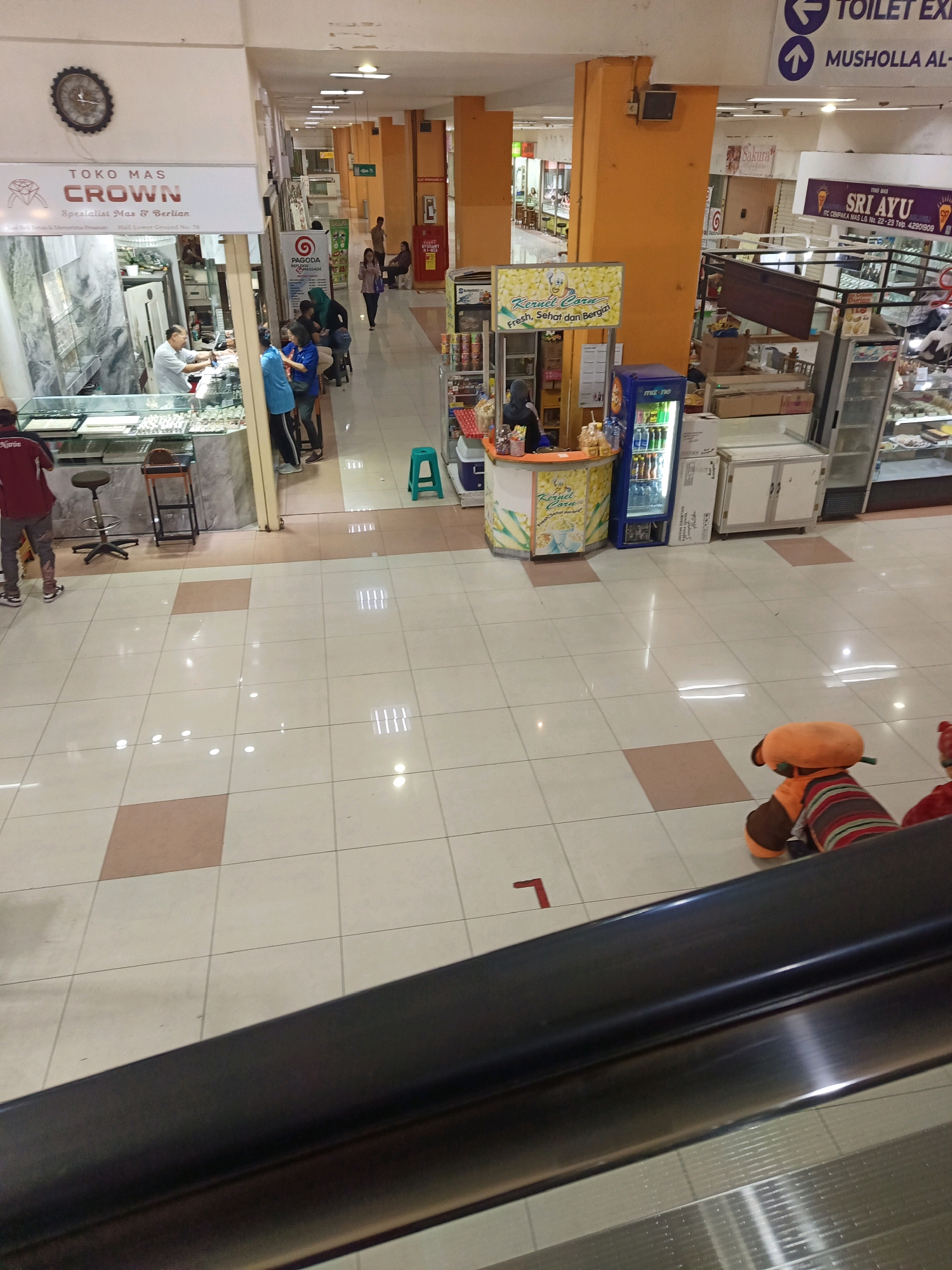 The image shows an interior view of a shopping mall in Central Jakarta, Indonesia. In the foreground, a dark escalator railing is visible, suggesting the photo was taken from an elevated position, likely another escalator or a walkway. The floor is tiled with a light beige and subtle pink pattern.
To the left, a jewelry store named "TOKO MAS CROWN Spesialis Emas & Berlian" (Gold Shop Crown Specialist Gold & Diamonds) is prominent. Inside the store, a person in a white shirt is behind a counter, and display cases filled with jewelry are visible. A customer in a red shirt stands outside the counter. Two bar stools are placed near the store's entrance.
Further down the mall corridor, several stalls and shops are present. One stall features a colorful advertisement for "Kernel Corn" with images of popcorn. Adjacent to it is a blue refrigerator unit. In the background, people are seen walking and standing. Signs indicating "TOILET EXIT" and "MUSHOLLA AL-" (likely referring to a prayer room) are visible on the ceiling. Another shop sign reads "SRI AYU" with some text below it.
In the mid-ground, a plush toy in an orange and striped outfit sits on the floor, facing away from the camera. The mall environment appears to be clean and well-lit, with overhead lighting fixtures visible. The overall impression is that of a typical indoor shopping mall environment.
