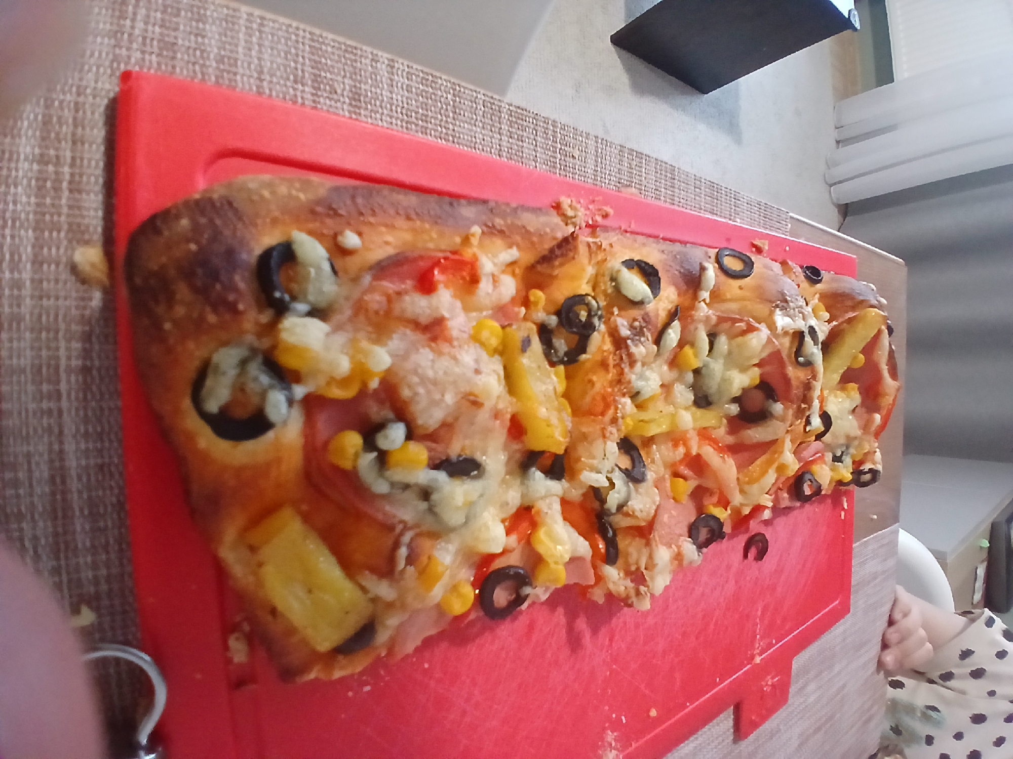 The image shows a long, rectangular pizza topped with ham, corn, olives, and cheese, placed on a red cutting board. The cutting board is on a woven placemat, which is on a table. In the background, there is a window with white curtains and a person's hand wearing a polka-dot shirt. The scene appears to be indoors, possibly a home kitchen or dining area. The lighting is natural and soft. There are no identifiable landmarks or details that would indicate a specific location within Yekaterinburg, Russia.