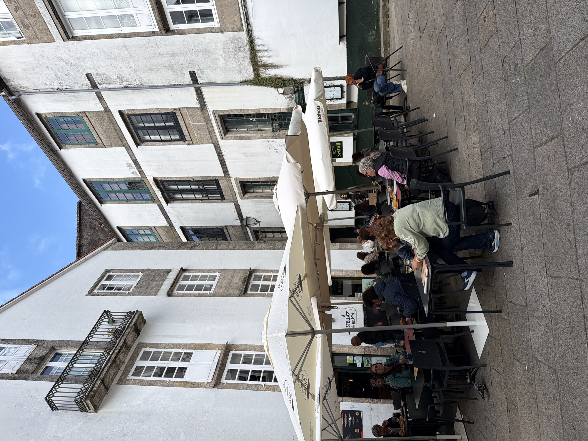 The image shows an outdoor cafe scene in Santiago De Compostela, Spain. Tables and chairs are arranged on a stone-paved area, shaded by large white umbrellas. People are seated at the tables, some eating, drinking, or talking. Buildings with white facades and various window styles form the backdrop. Some windows have shutters, and a balcony is visible on one of the buildings. A few individuals are walking along the street.