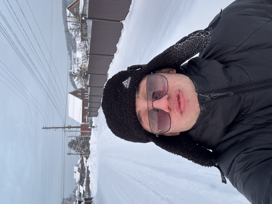 A selfie-style portrait depicts a young male centered in a snow-covered street in Ольховка, Russia, under an overcast sky. The subject wears a black Adidas ushanka-style winter hat, dark-framed sunglasses with purple-tinted lenses, and a black puffer jacket. His lips are puckered, appearing chapped. The background features a snow-covered road with visible tire tracks, flanked by snowbanks. To the left, multiple wooden houses with snow-laden roofs are visible. To the right, a dark brown corrugated metal fence runs alongside the road, beyond which another house with a steep, snow-covered brown roof is present. Utility poles and overhead power lines traverse the upper portion of the scene. In the distance, snow-covered hills and evergreen trees complete the wintry landscape.
