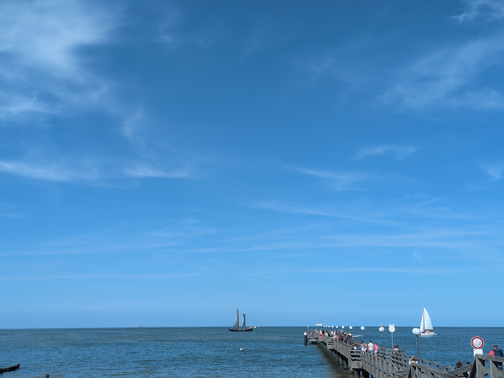 The image depicts a sunny day at the pier in Kühlungsborn, Germany. The sky is a vibrant blue with scattered white clouds, and the sea is calm and blue. A wooden pier extends from the right side of the frame into the sea, with many people walking along it. The pier is lined with lampposts and railings. Two sailboats are visible in the distance on the water. One is a large, dark-sailed schooner, and the other is a smaller white sailboat. The scene captures a typical summer day at a seaside resort.