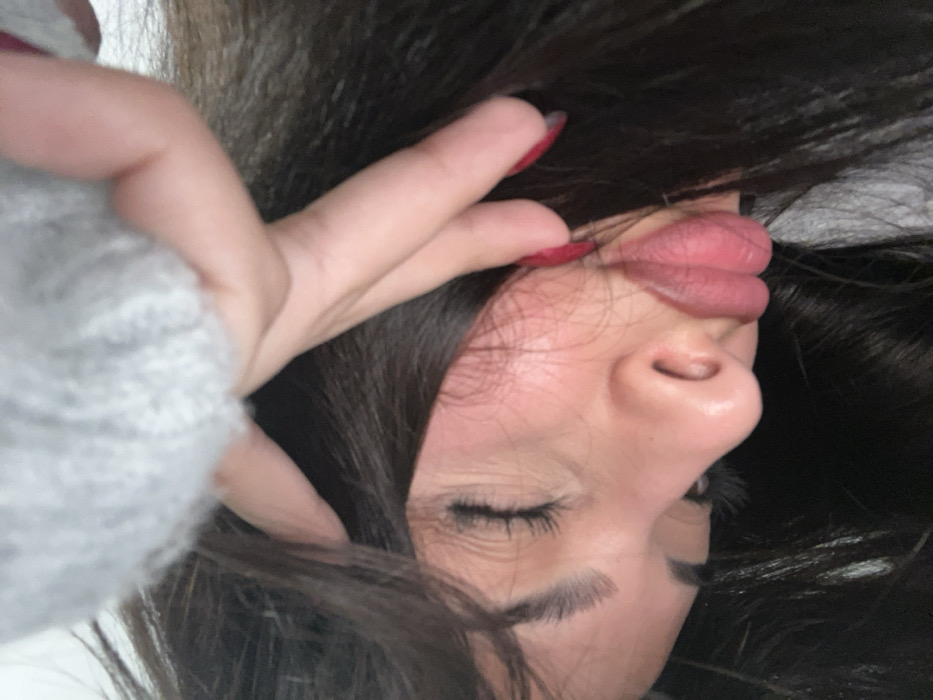 A close-up, angled portrait features an individual with long, dark hair, positioned with their head tilted. Their eyes are closed, or looking downwards, and their lips are distinctly pursed outwards. A hand, adorned with red nail polish, is visible in the upper portion of the frame, touching the subject's hair and head. A portion of a grey, textured, possibly fuzzy garment is visible, suggesting it is worn by the individual. The background is out of focus, providing no discernible environmental details. The image exclusively depicts a close-up portrait and offers no visual information to determine a specific location within Tbilisi, Georgia, or any other city.
