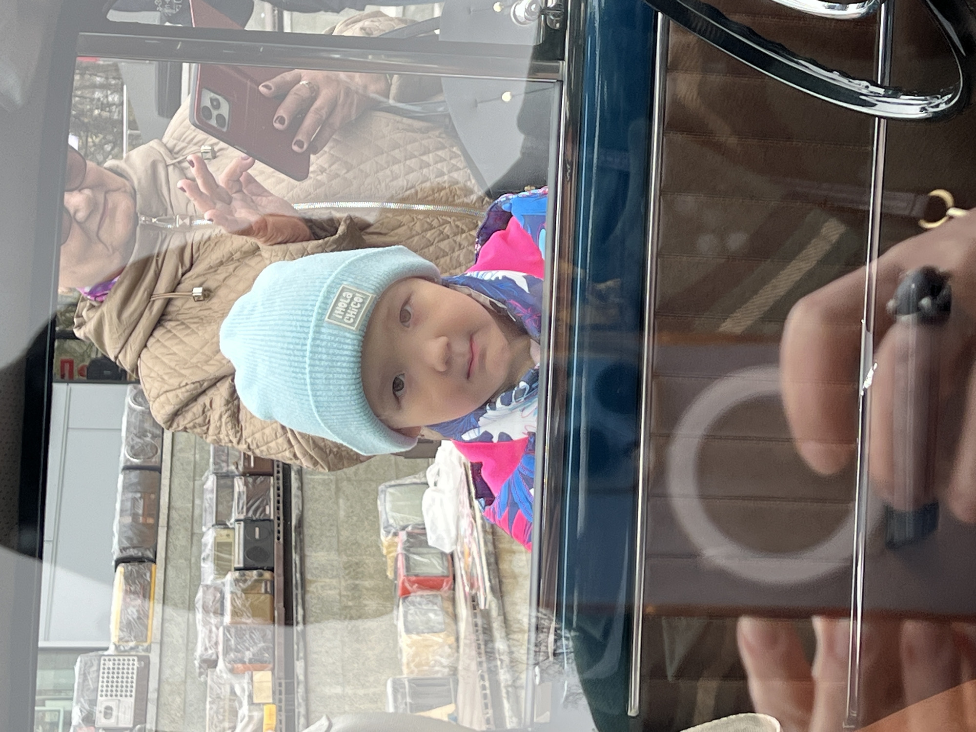 The image depicts a reflection in a vehicle window, likely a car. Visible within the reflection are: a young child wearing a light blue beanie with visible text, a beige quilted jacket, and a colorful shirt; an older woman holding a mobile phone, potentially taking a photo of the child; a display of radios in plastic packaging and on shelves visible through the window. The background contains elements suggesting the Saint Petersburg, Russia as the radios displayed in plastic packaging are commonly sold in the city. The vehicle’s interior includes a steering wheel and a wooden panel. Part of an arm and hand holding the mobile phone are reflected in the window as well.