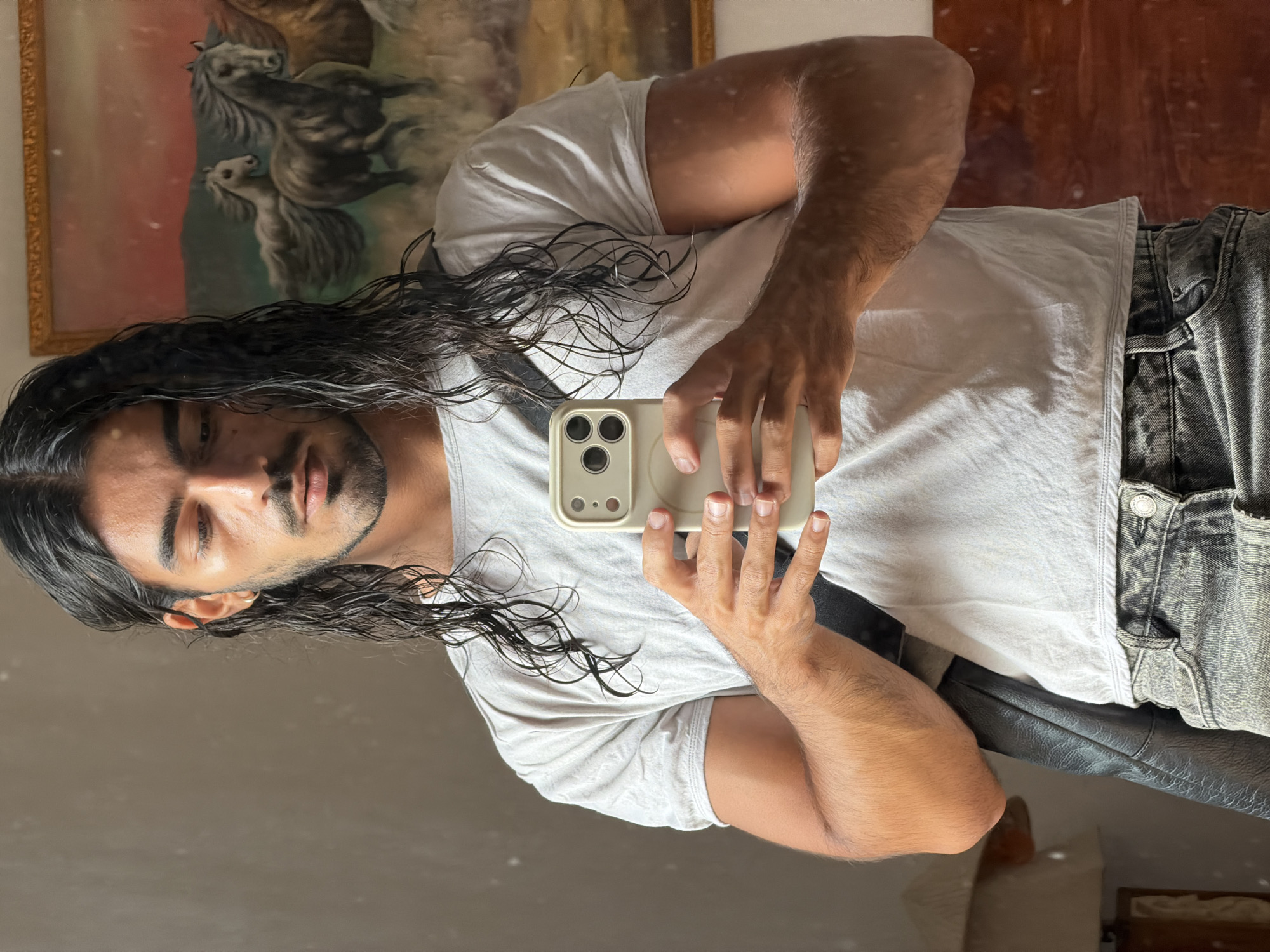 A man with long, dark, wet hair is taking a selfie in a mirror. He is holding an iPhone in a beige case. He is wearing a white t-shirt and grey denim pants. Behind him, a painting of horses is visible. He is located in North Kuta, Indonesia.