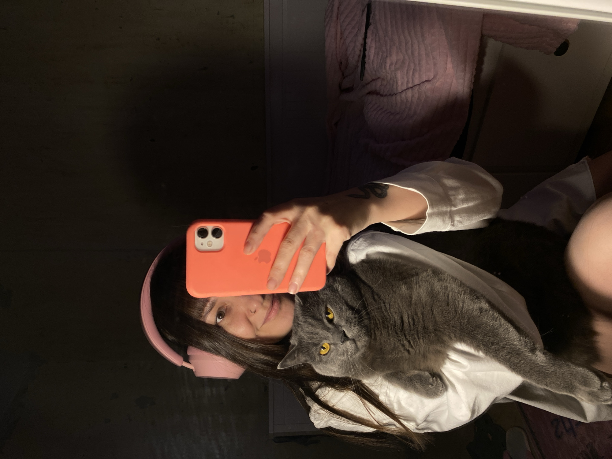 A woman is taking a selfie with a gray cat. She is holding an orange phone in her right hand. She is wearing pink headphones. The background shows a dark wall, a light-colored piece of furniture, and a pink blanket. Location details are unavailable.