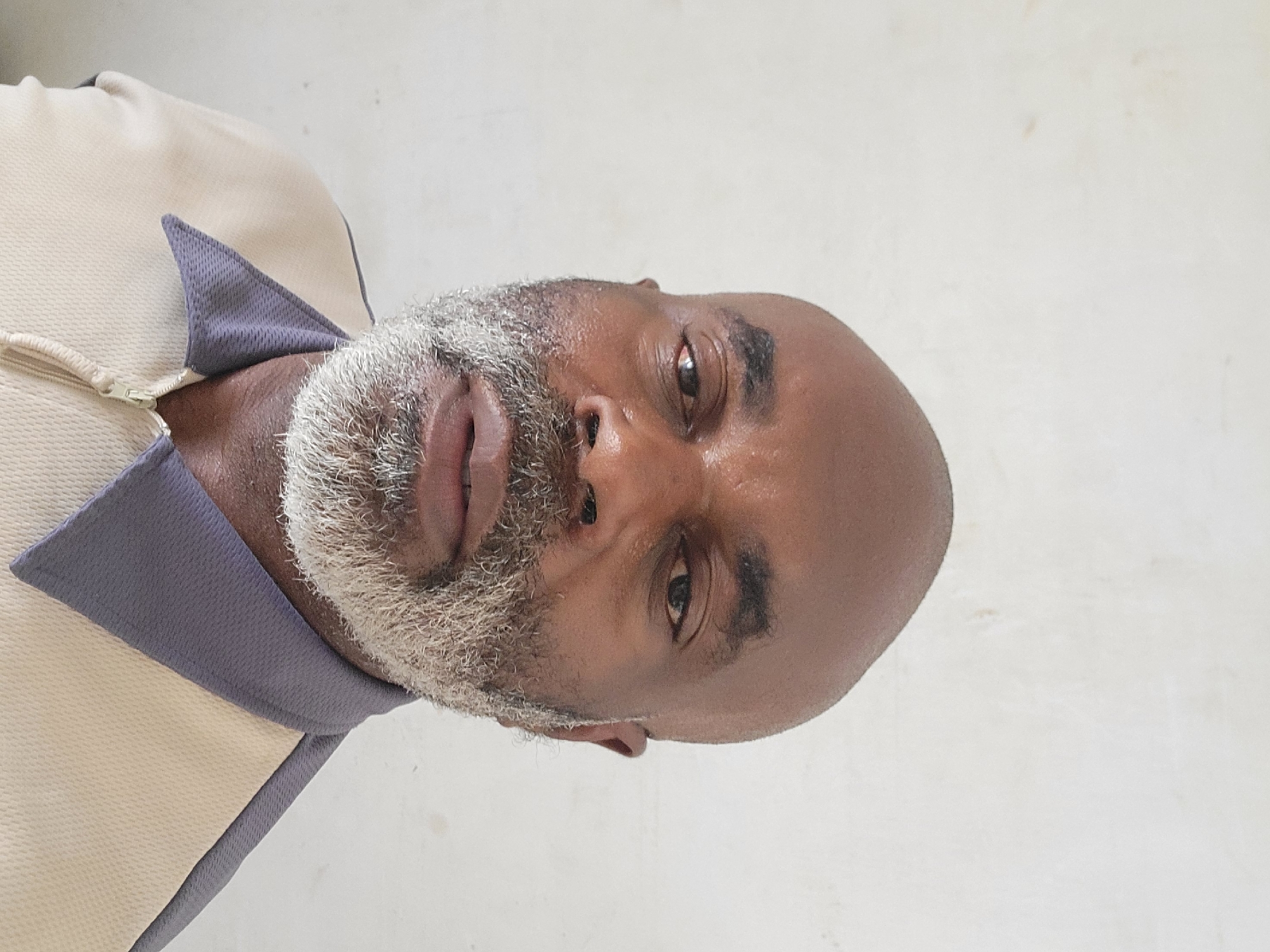 A rotated, close-up portrait features a dark-skinned man, potentially located in Maiburiji, Nigeria, looking directly at the viewer. He has a full, neatly trimmed beard that is predominantly grey or white, contrasted with his dark skin. His scalp appears bald or has very short dark hair. His eyes are open and focused, and his expression is neutral, possibly thoughtful or serious. There is a slight sheen on his skin, particularly on his forehead and around his eyes, which could be due to lighting or natural oils.
The man is wearing a collared polo shirt. The main body of the shirt is a light cream or beige color with a textured, possibly ribbed fabric. The collar and triangular accents on the shoulders are a contrasting grey-purple color, appearing to have a mesh-like texture. A partial zipper is visible at the neckline of the shirt. The background is a plain, light off-white or white surface, suggesting an indoor setting or a simple backdrop. No specific location cues or visible text are present within the image itself. The time of day and weather are not discernible.