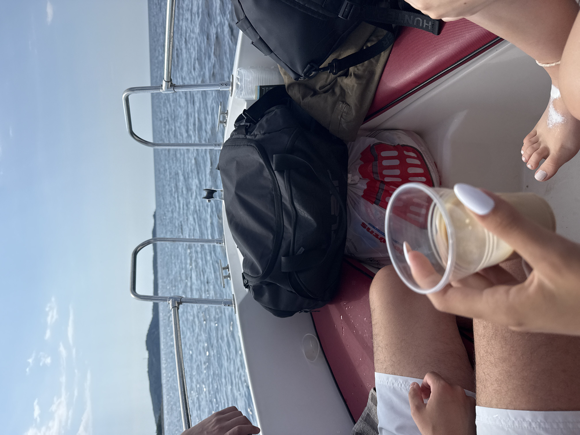 An outdoor, first-person perspective shot shows two pairs of legs and a hand holding a drink aboard a boat sailing on the Aegean Sea near Potos, Greece. The boat's white hull and red seating are visible, along with two black duffel bags and a pair of green trousers. The sea is calm, and a hilly, tree-covered island is visible in the distance under a clear blue sky with sparse white clouds. The hand holding the drink has white-painted fingernails. The legs are tanned and one pair is wearing white shorts. There is sunscreen on one of the feet. The boat appears to be moving at a moderate speed.