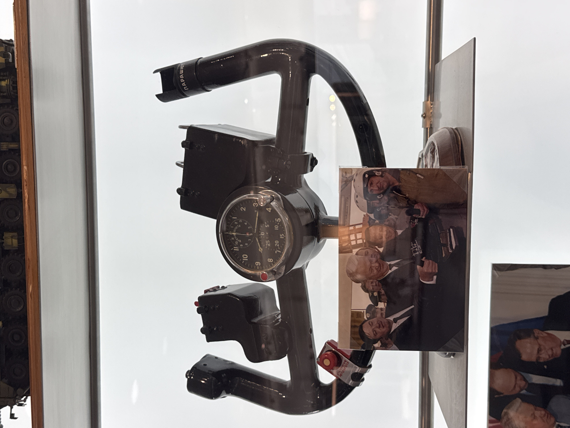 This is a close-up, vertically oriented, eye-level photograph taken through glass, showcasing a black, complex aircraft control column. The control column features a circular dial with numbered markings and hands, likely an altimeter or similar instrument. Attached to the column are several black boxes and levers, some with red buttons. Text visible on the control column includes "ПАРАШЮТ" and "ОТКЛЮЧЕНИ АВТОПИЛОТА" in Cyrillic, suggesting it is part of a Soviet or Russian aircraft's autopilot system.
To the right of the control column, several photographs are displayed, also behind glass. These photographs show groups of men in formal attire, mostly suits and some military caps. The men appear to be smiling and engaged in conversation, suggesting a celebratory or formal gathering. One of the photos shows a man in a pilot's helmet. The overall setting appears to be a museum exhibit or a display case, given the reflective glass and the context of historical artifacts. The lighting is bright and diffused, creating reflections on the glass surfaces. The background is predominantly white, suggesting a clean and well-lit display environment. The presence of Cyrillic text and the design of the control column strongly suggest a Russian context, possibly related to aviation history.