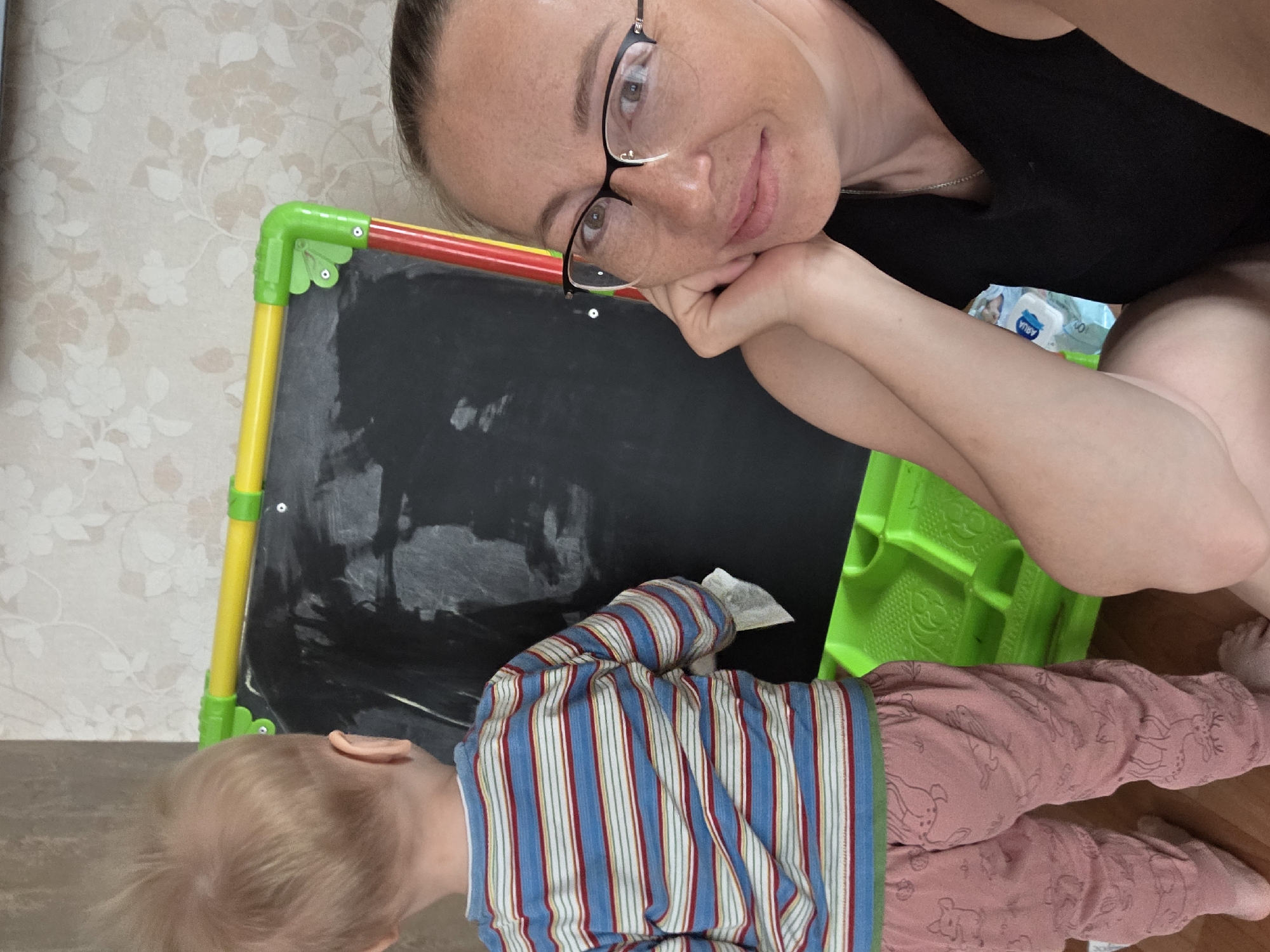This indoor image captures a woman and a young child in what appears to be a home setting. The woman, positioned in the upper part of the frame, has light brown hair pulled back, wears black-framed glasses, and a black top. She looks directly at the camera with a gentle, slightly smiling expression, resting her chin on her hand.
In the foreground, a young child, seen from behind, is focused on a child's blackboard. The child has blond hair and is dressed in a long-sleeved shirt with vertical blue, red, and white stripes, and light pink patterned pants. They appear to be engaged with the blackboard, possibly drawing or cleaning it with a small white object in their hand.
The blackboard has a vibrant green plastic frame and colorful yellow and red supports. It shows faint white chalk marks on its black surface. In the background, behind the woman, a white and blue package with the text "AQUA" is visible, suggesting it might be a pack of wipes. The room features light-colored wallpaper with a subtle floral pattern and wooden floorboards. The scene conveys a calm, intimate moment, likely a parent observing a child at play.