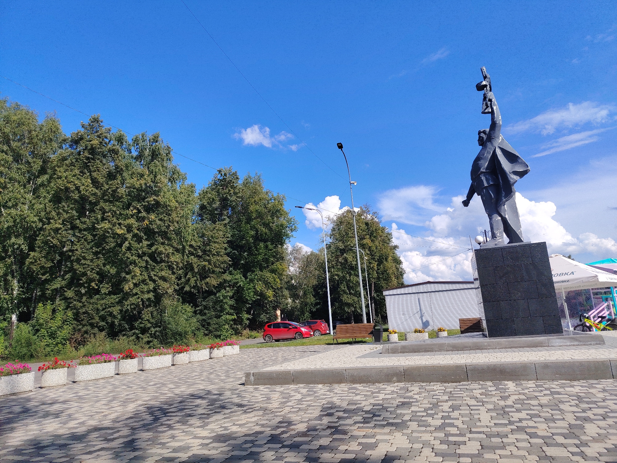 The image shows a view of a park area with a monument in Kemerovo, Russia. In the foreground, there is a paved area with a repeating pattern of grey and black cobblestones. Along the left side of the paved area, there is a row of rectangular planters filled with pink and red flowers. Further back, a red hatchback car is parked on a grassy area. Behind the car, a line of trees forms a dense green backdrop. To the right of the car, there is a wooden park bench and a lamppost. Dominating the right side of the image is a tall, dark-colored monument, which appears to be a statue of a person raising an object above their head, standing on a square base. Beyond the monument, a corrugated metal building and a white tent with text on it are visible. The sky is a clear blue with scattered white clouds.
