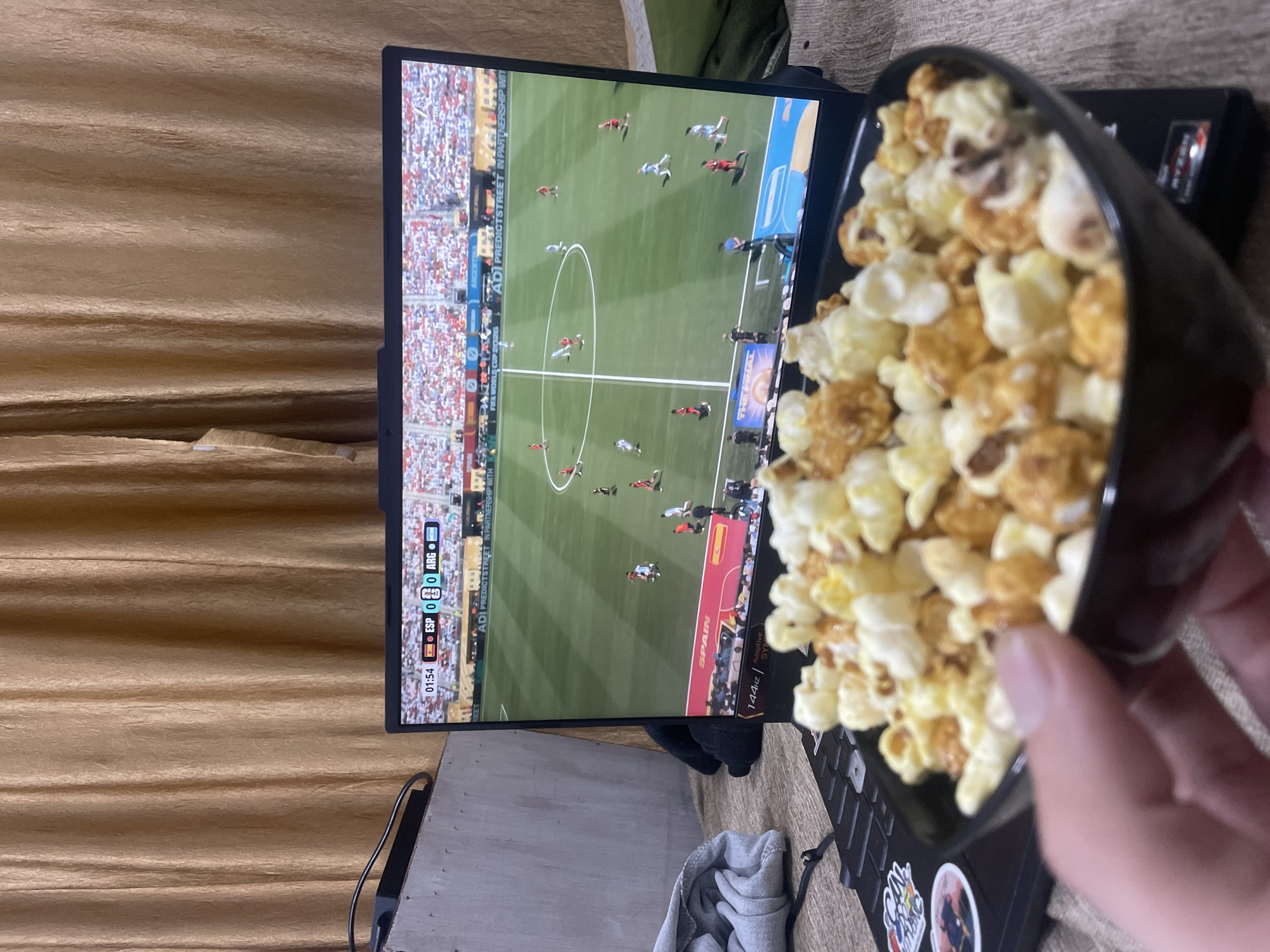 A bowl of popcorn is held in the foreground of the image, with a person's thumb and finger visible at the left edge of the frame. Behind the popcorn, a laptop is open, displaying a soccer match. The laptop screen shows players on a green field with a stadium full of spectators in the background. A score graphic indicates "ESP 0 0 ARG" and a timer shows "01:54". The laptop is positioned on a surface that appears to be a bed or couch, covered with a patterned blanket. To the left of the laptop, a grey cloth is draped over an object, possibly clothing, and a black cable is connected to the laptop. The background of the scene consists of a golden-brown curtain. There is no discernible connection to the city of Bhagsu, India, in the provided image.