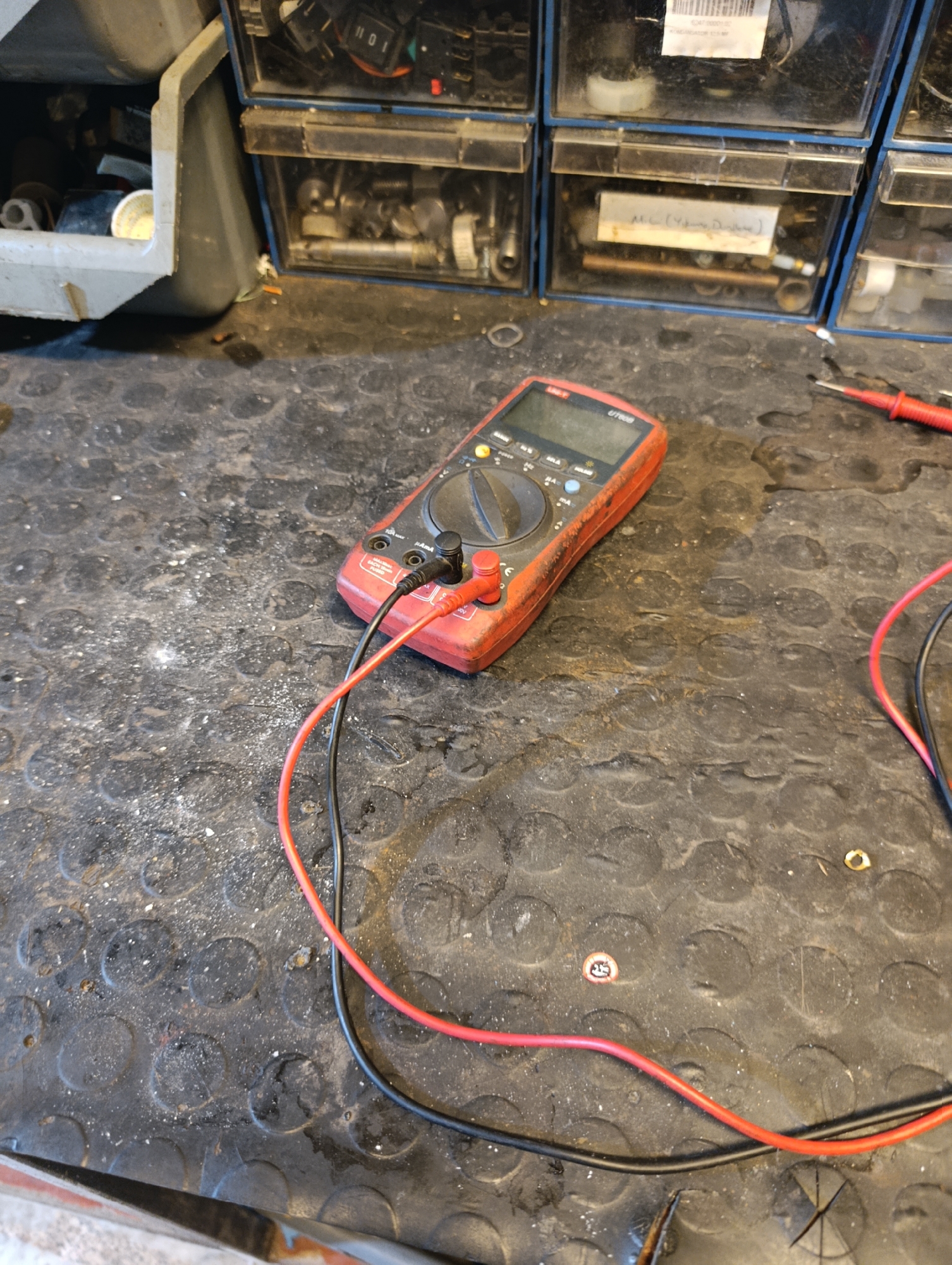 The image shows a red digital multimeter on a dirty workbench. The multimeter has black and red test leads connected to it. The workbench is made of a dark, textured material and is littered with dust and debris. In the background, there are several drawers or compartments filled with various tools and small parts. The overall scene suggests a workshop or repair environment. The location is specified as Ümraniye, Turkey.