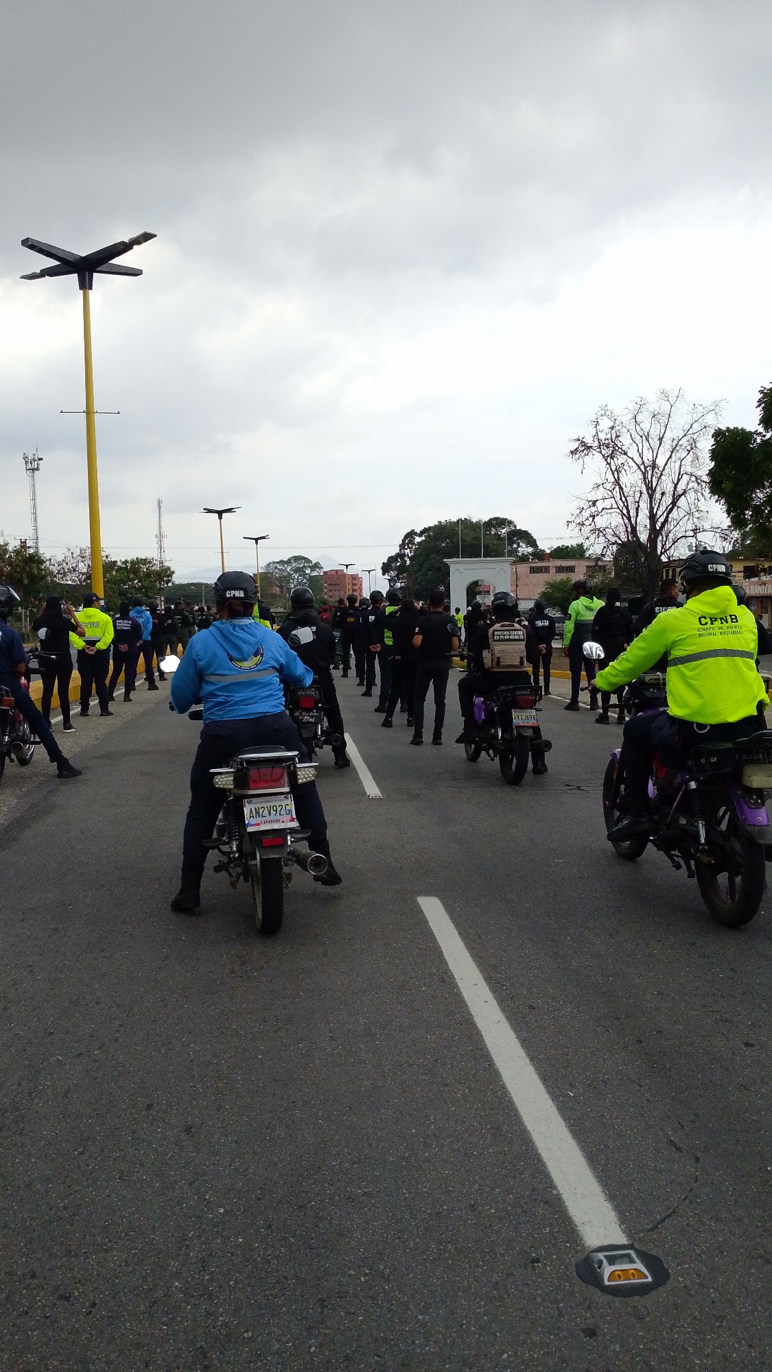 The image depicts a street scene in Tinaquillo, Venezuela. A group of individuals, identified as "CPNB" (Cuerpo de Policía Nacional Bolivariana) police officers, are present, some standing and others on motorcycles. The officers are wearing uniforms, with some sporting neon green jackets. The motorcycles are parked on the asphalt road, with a white lane marking visible. In the background, there are buildings, trees, and distinct streetlights. The sky is overcast, suggesting cloudy weather conditions. License plates are visible on some of the motorcycles, notably "AH2Y92G".