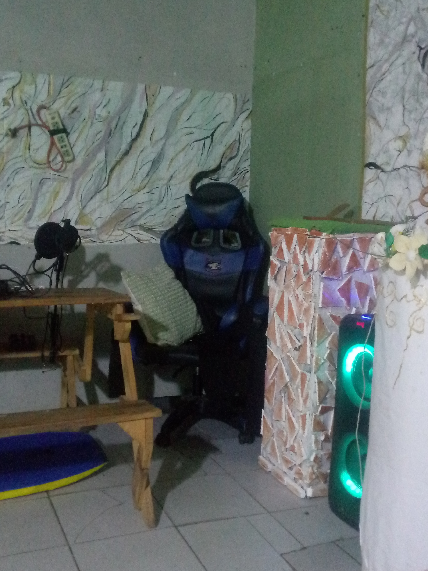 The image shows an interior scene, possibly within a home or studio, in Juan Griego, Venezuela.
A blue and black gaming chair with a green pillow rests in the center of the room. To its left is a wooden table holding a microphone with a pop filter. Underneath the table, there is a blue and yellow cushion.
To the right of the chair, a decorative structure with a mosaic-like pattern of red and white triangles stands next to a black speaker with green illuminated rings.
The walls in the background feature varied patterns. One section has a mural with what appear to be abstract white and gray lines with a power strip, while another section is a solid green.
The floor is covered with white tiles.