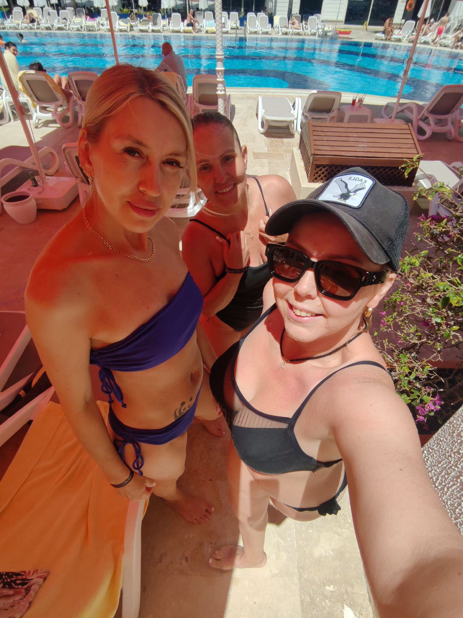 Three women are posing for a selfie near a swimming pool. The woman in the foreground is wearing a black bikini, sunglasses, and a black baseball cap with a white eagle on the front. To her left, a woman is wearing a blue bikini. Behind them, the third woman is wearing a black swimsuit. The pool is surrounded by lounge chairs and umbrellas. The photo was taken in Alanya, Turkey.