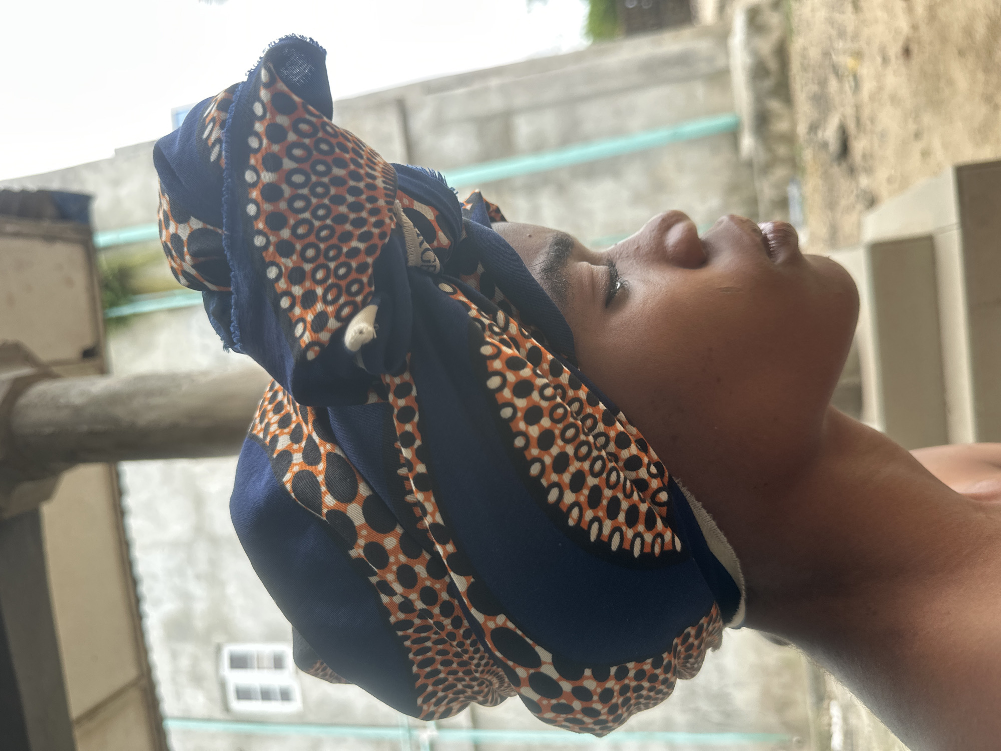A close-up, profile shot shows a person wearing a blue headwrap with an orange and white circular pattern. The person's face is turned to the right, with their eyes closed and lips slightly parted. The background is blurred, but appears to be an outdoor setting with a building and some vegetation. The scene is set in the city of Badagry, Nigeria.