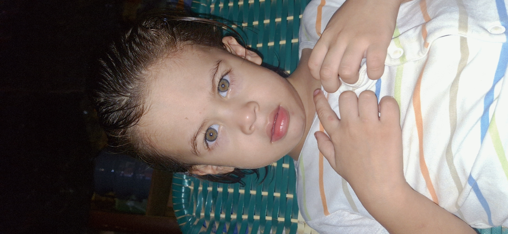 A close-up, eye-level, vertical shot shows a young child with wet, dark hair looking directly at the camera. The child has light skin, hazel eyes, and slightly plump lips with a touch of pink. The child is wearing a white collared shirt with thin horizontal stripes in orange, light brown, green, and blue. The child's hands are crossed over their chest. Behind the child, a green, woven chair is visible, and the background is dark and out of focus, suggesting an indoor setting at night. The image was taken in Vallecito, Venezuela.