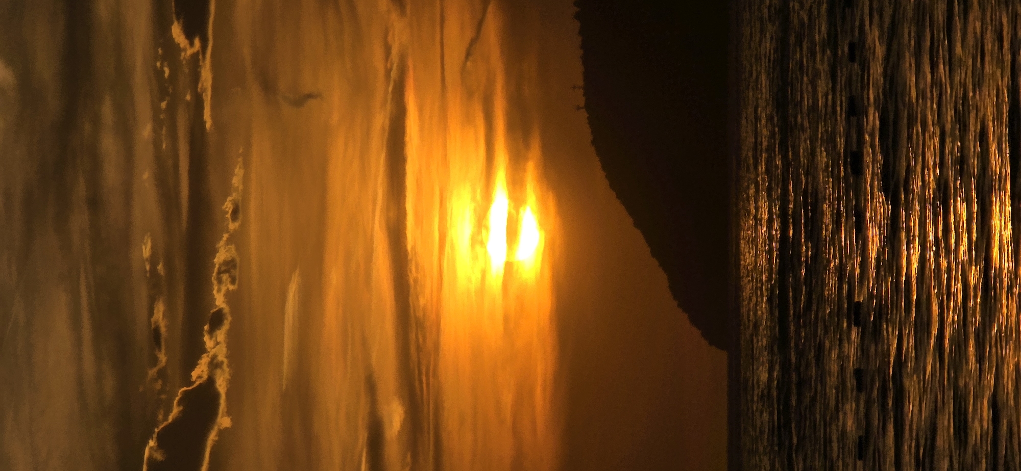 The image depicts a sunset reflected in the water near Bar, Montenegro. The left side shows the rippling surface of the water illuminated by the setting sun, with the bright light creating streaks and patterns. A dark landmass is visible in the middle ground, partially obscuring the horizon. To the right, the water's surface appears textured, with long, vertical lines of light mirroring a nearby structure like a pier or a line of moored boats. The overall color palette is dominated by warm tones of orange and gold, transitioning to darker shades.